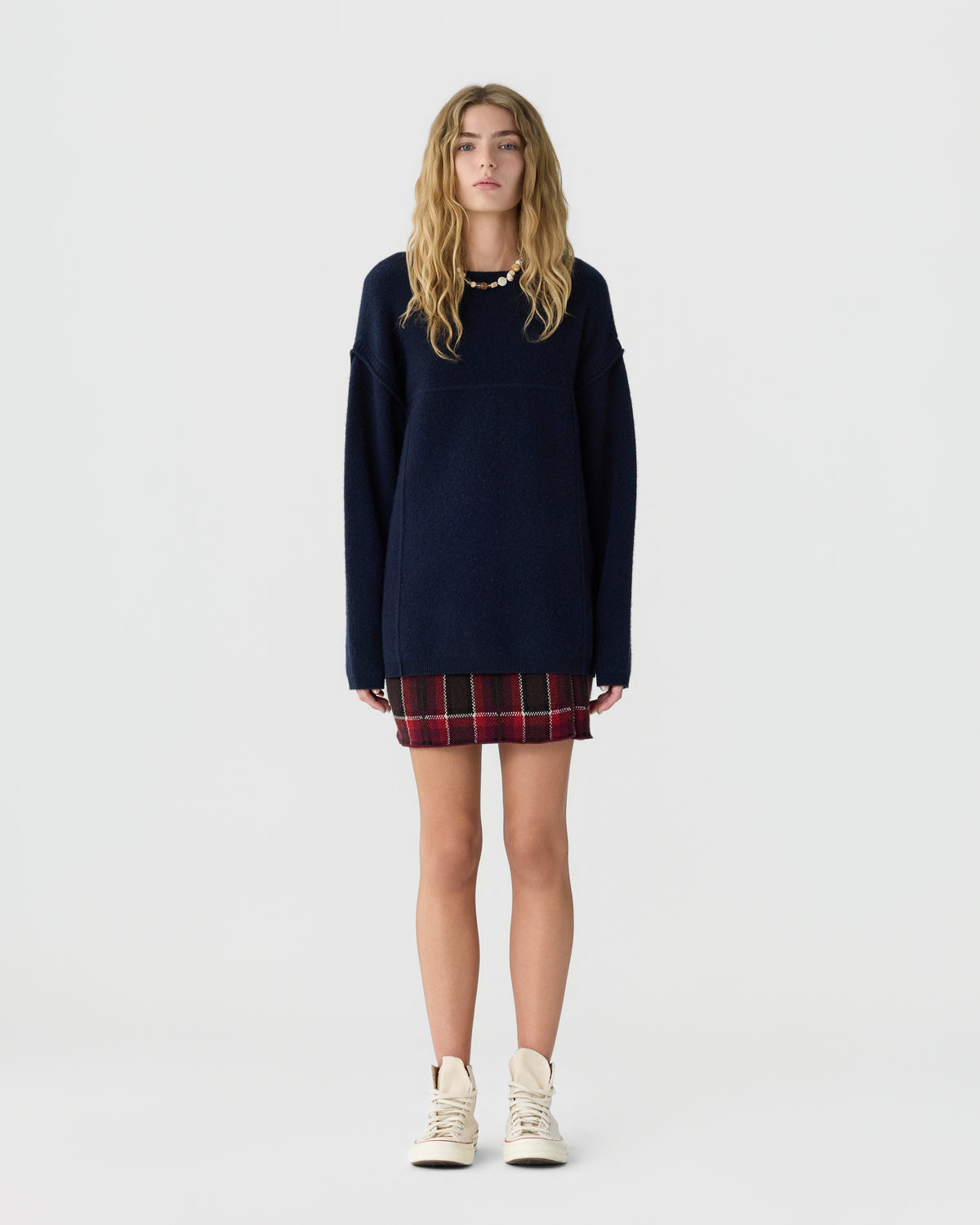 Woman wearing an oversized navy sweater with a red plaid skirt and cream sneakers, standing against a light gray studio background.
