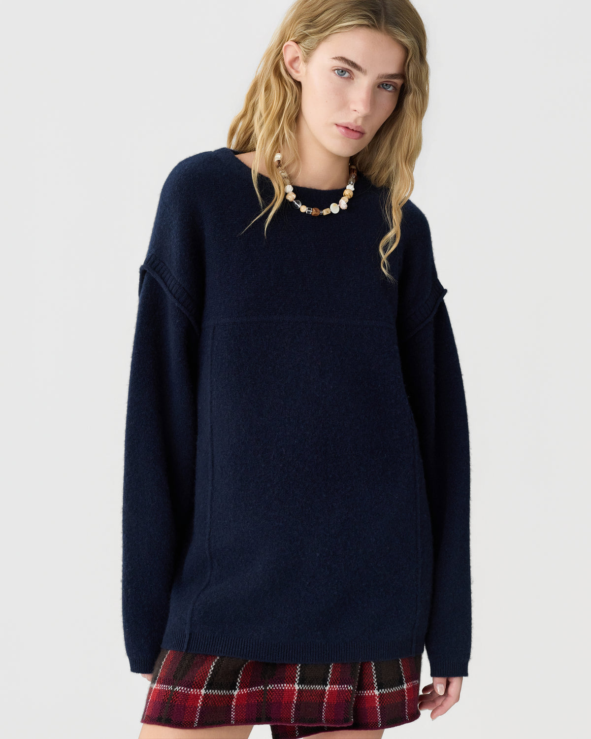 Model wearing a navy oversized sweater with a red plaid skirt and beaded necklace against a light gray background