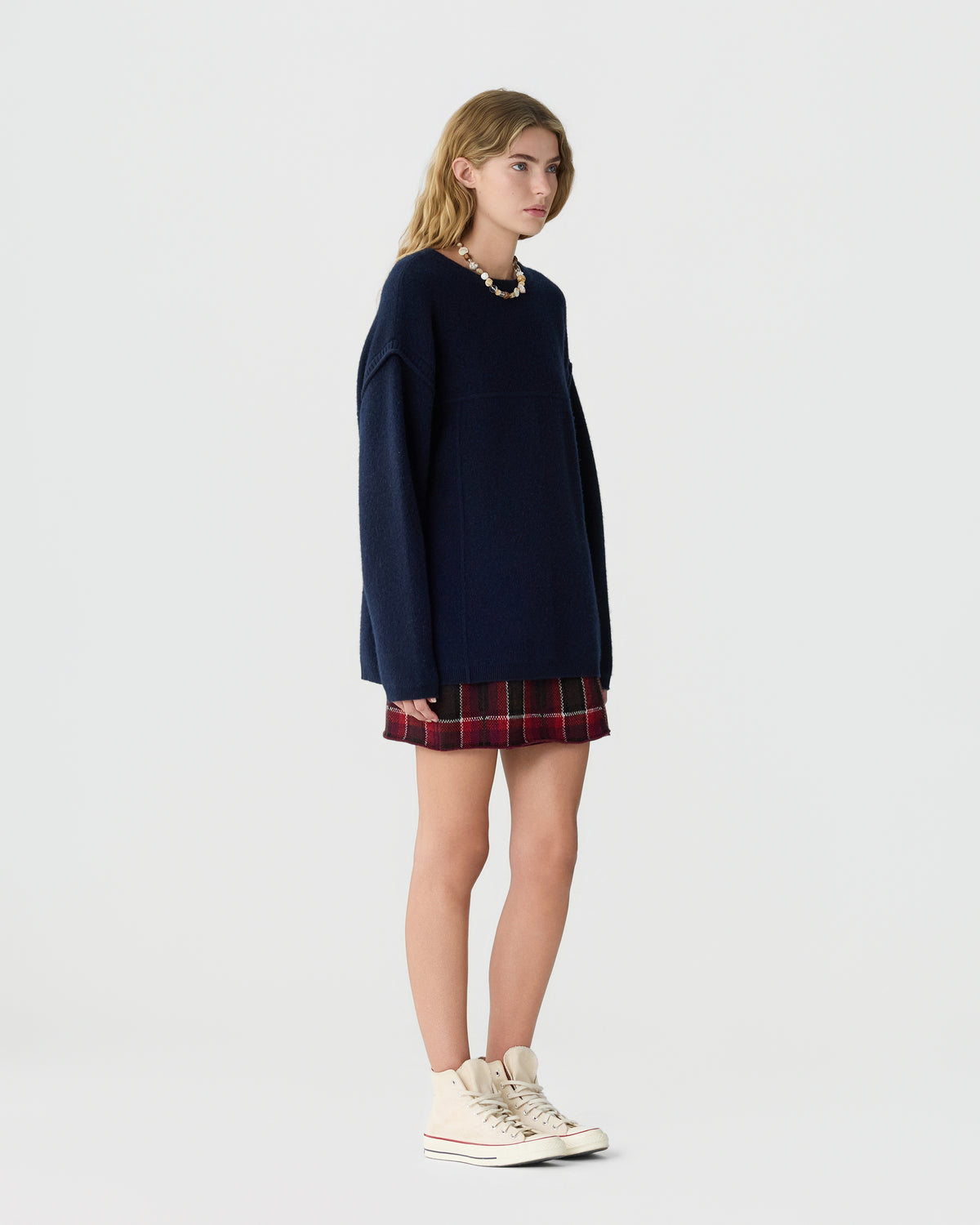 Woman wearing a navy long sleeve sweater over a red plaid skirt with white sneakers against a light gray studio background