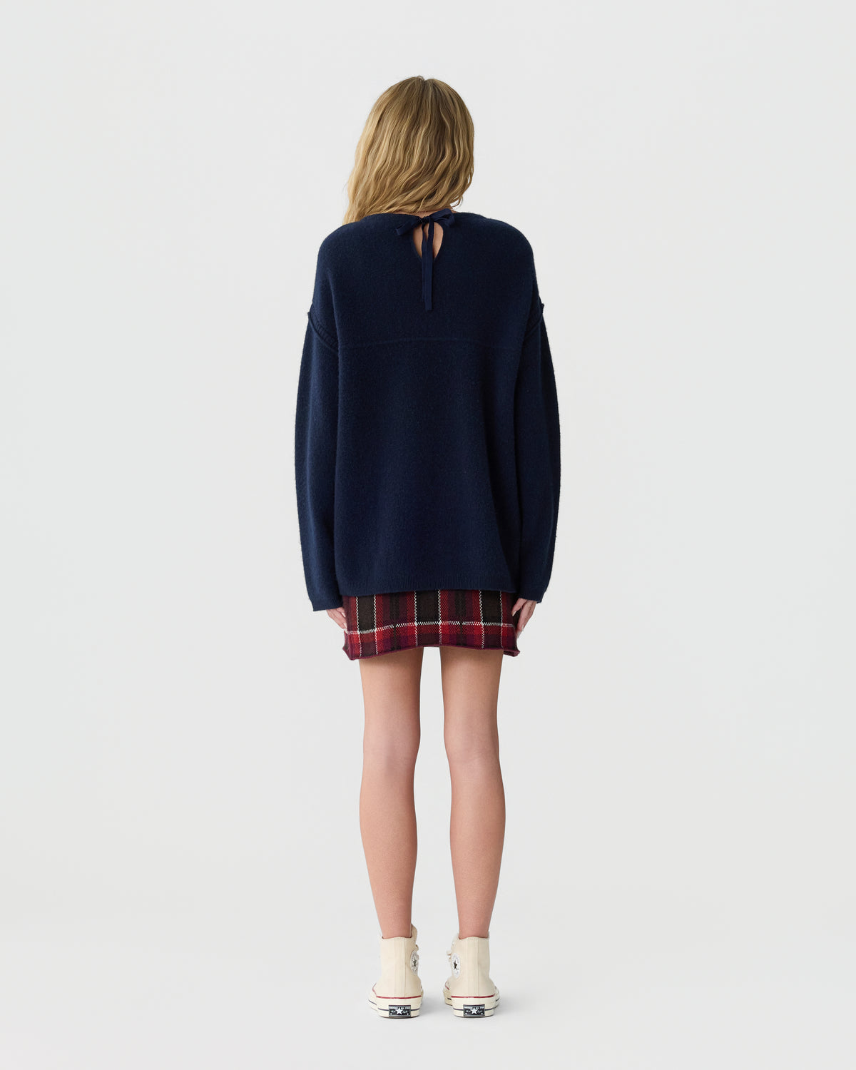 Rear view of a person wearing a navy sweater, red plaid skirt, white ankle socks, and low shoes against a light gray background