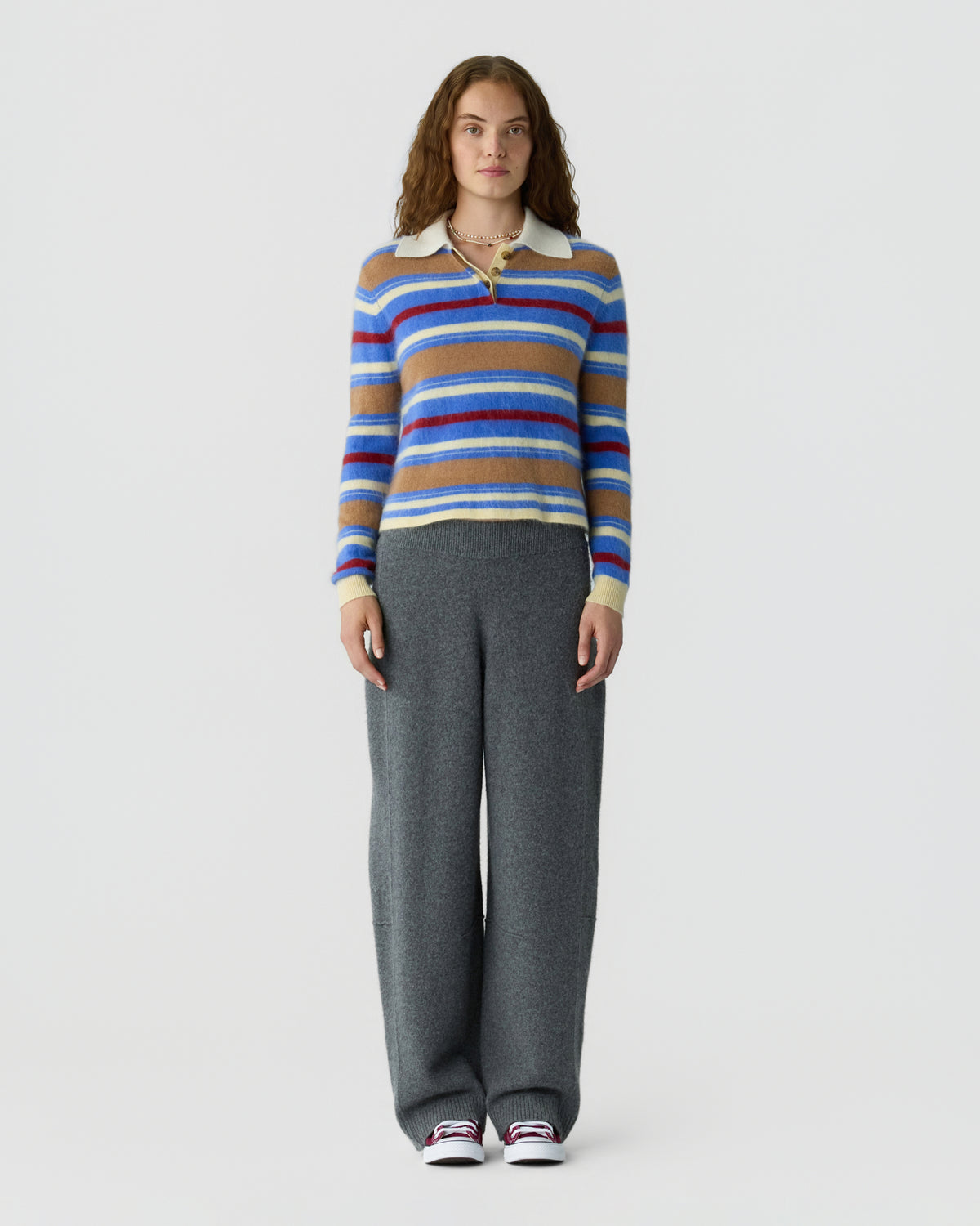Full length portrait of a woman wearing a blue red and tan striped collared sweater with wide leg gray trousers and white sneakers against a light background