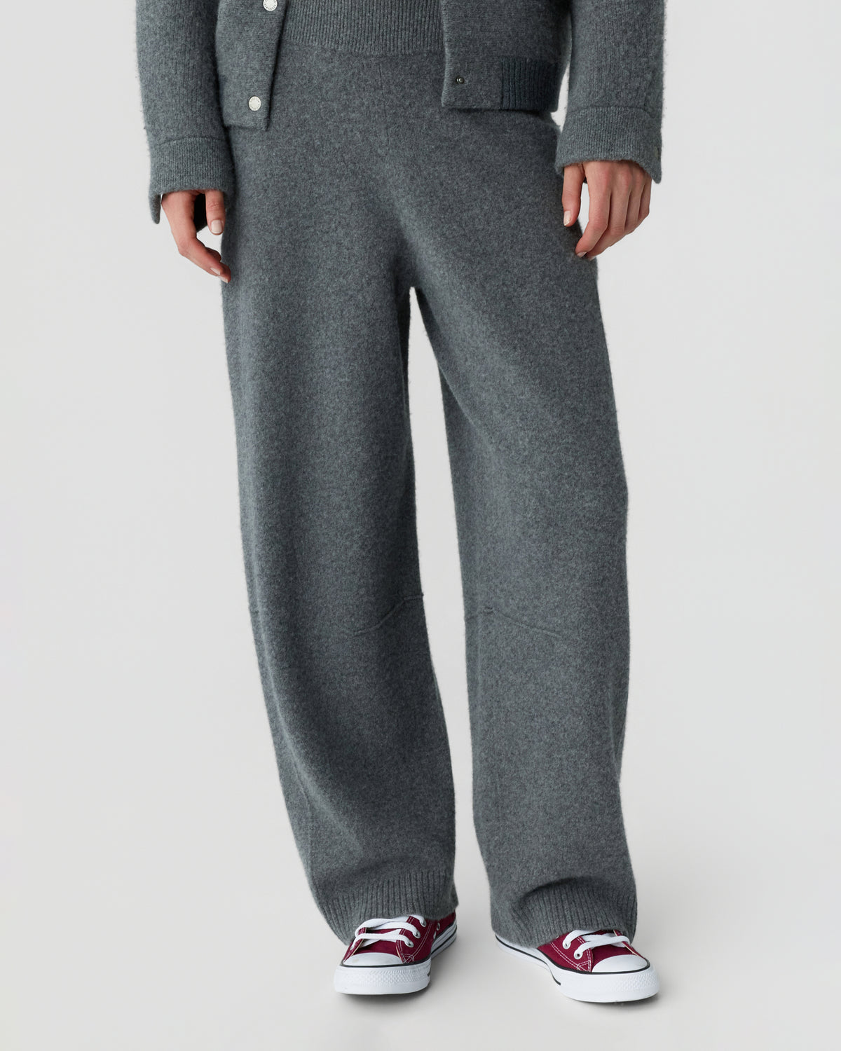 Model wearing wide leg charcoal knit trousers with a matching buttoned cardigan and burgundy canvas sneakers against a light gray background