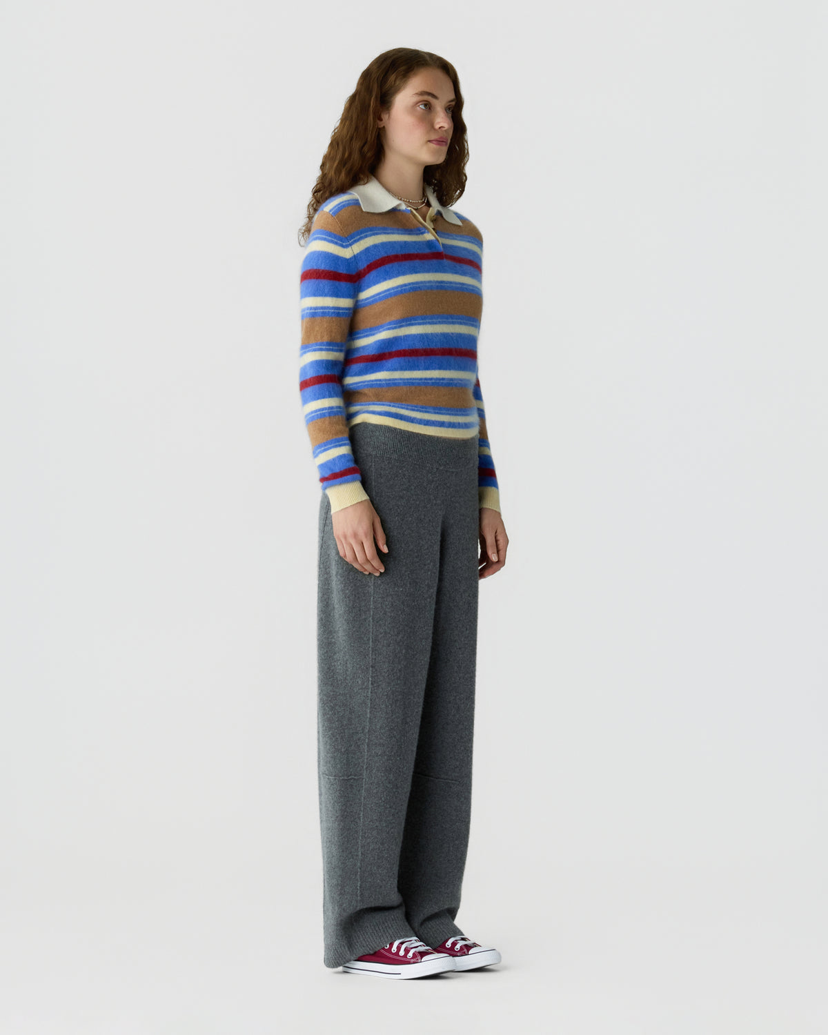 Full length view of a woman wearing a blue striped collared sweater with gray wide leg trousers and red and white sneakers against a light background