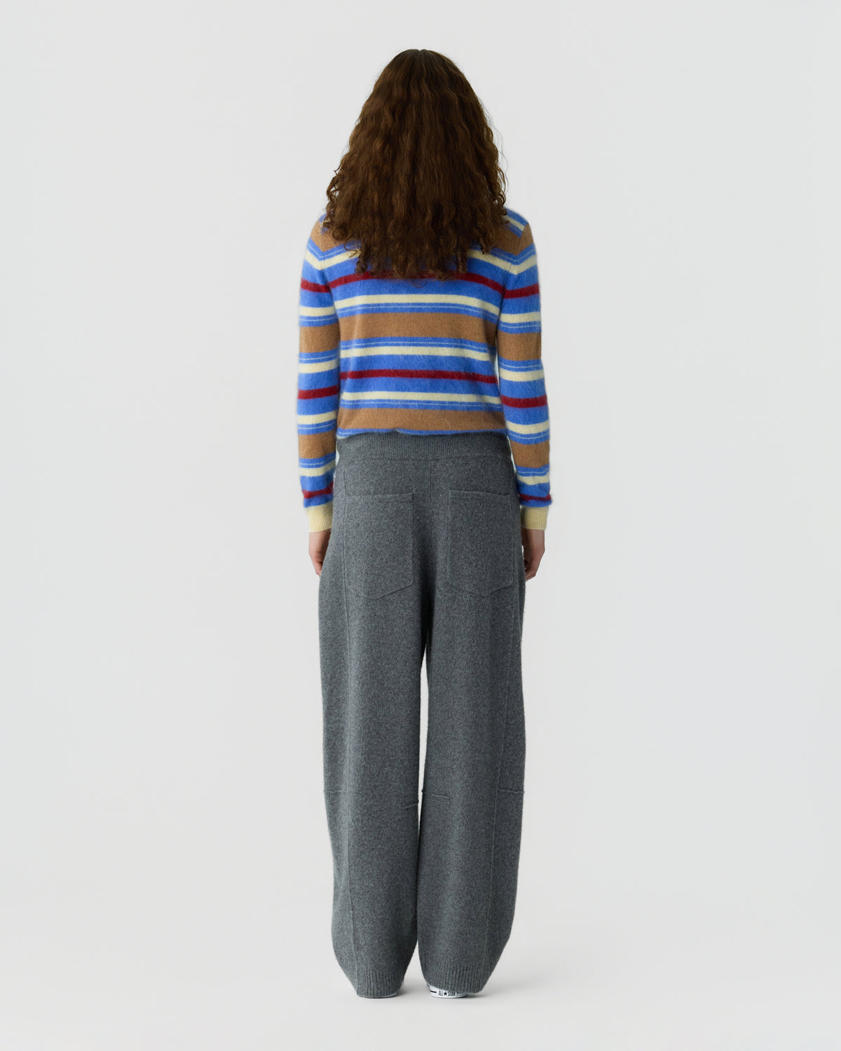 Rear view of a person wearing a multicolored striped long sleeve sweater with gray wide leg pants against a plain light background