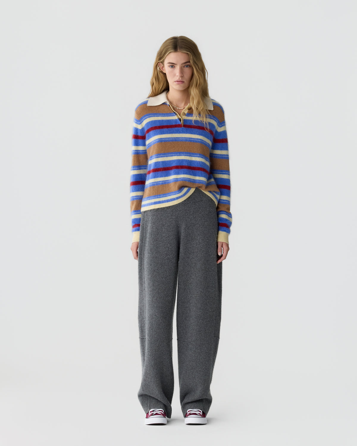 Full length view of a woman wearing a colorful striped sweater with a white collar, gray wide leg trousers, and red and white sneakers against a light background