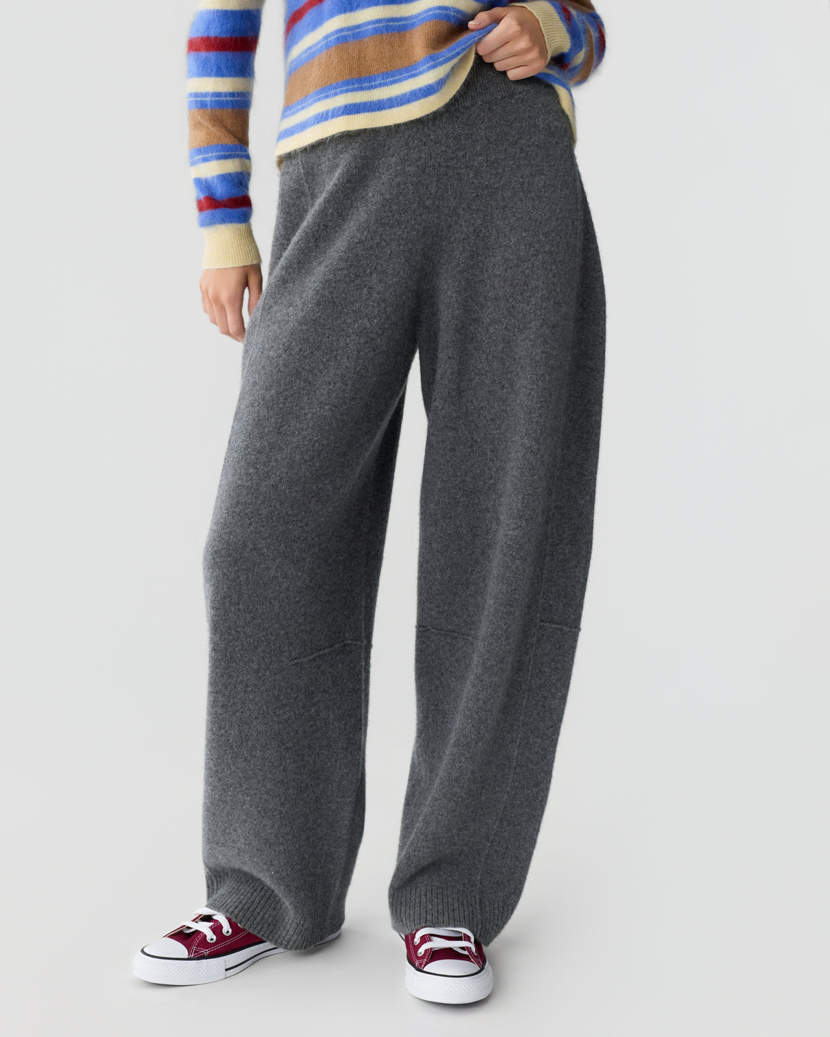 Cropped view of a person wearing wide leg charcoal gray trousers, a striped sweater, and red and white sneakers against a light gray background