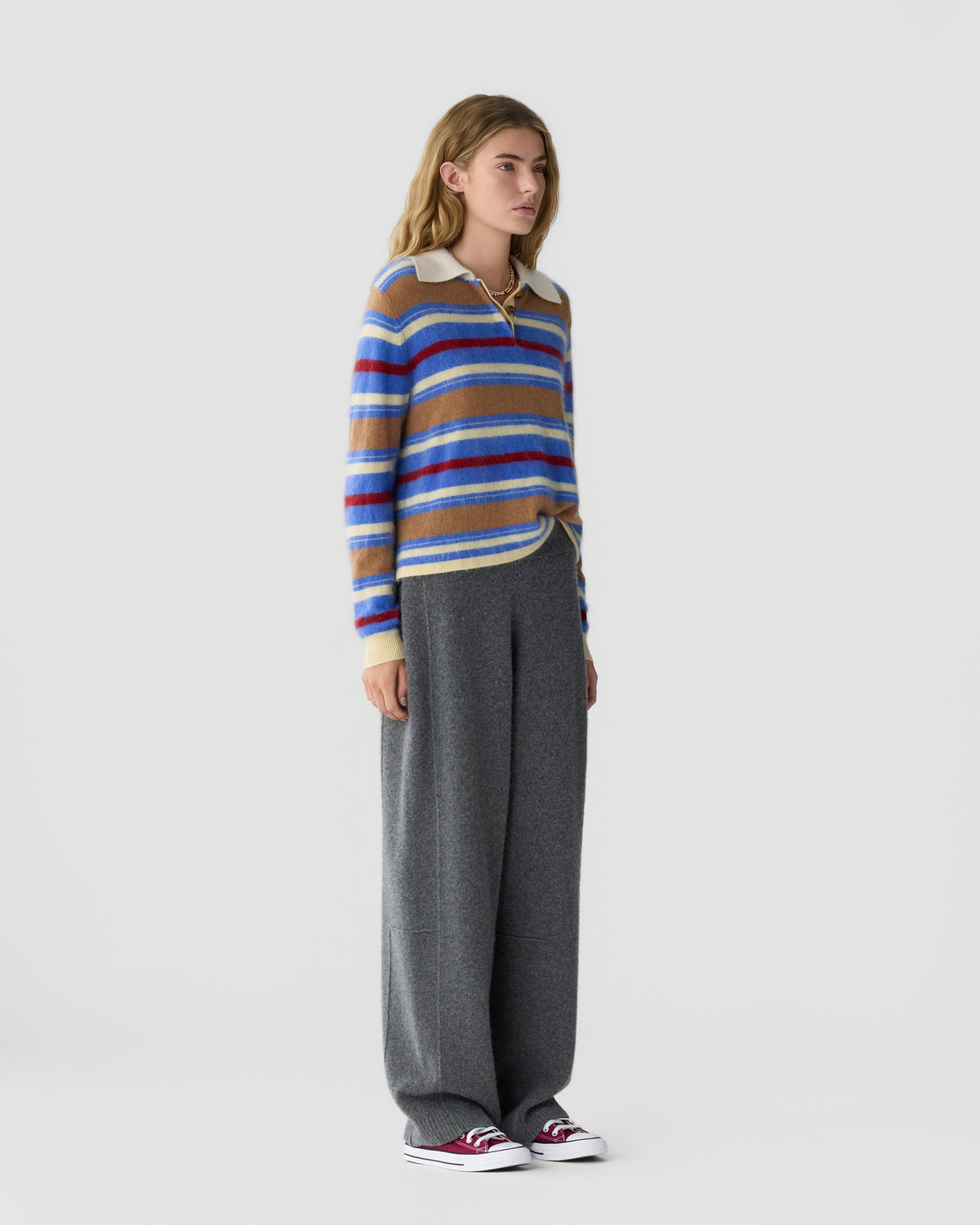 Woman standing in a studio wearing a blue, red, and tan striped sweater, loose gray trousers, and red and white sneakers against a light background