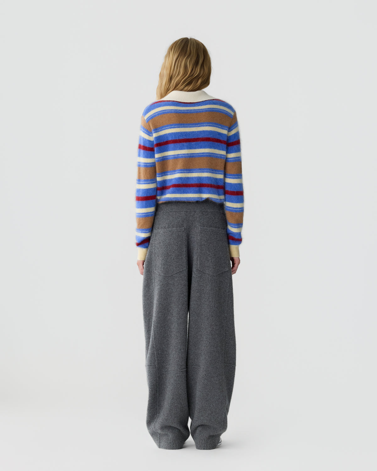 Model seen from behind wearing a colorful striped sweater and wide leg gray trousers against a plain light background