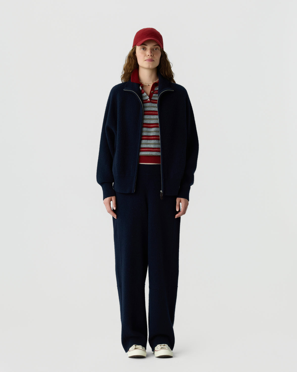 Full length person wearing a navy zip jacket and matching trousers with a striped shirt red cap and white sneakers against a light background