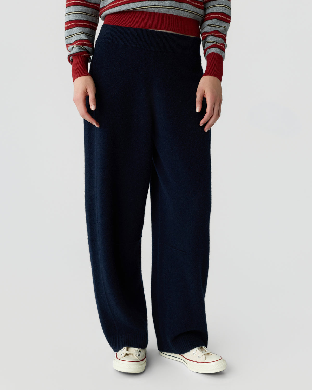 Cropped view of a person wearing a striped red gray and navy sweater with wide leg navy trousers and white low top sneakers against a light background