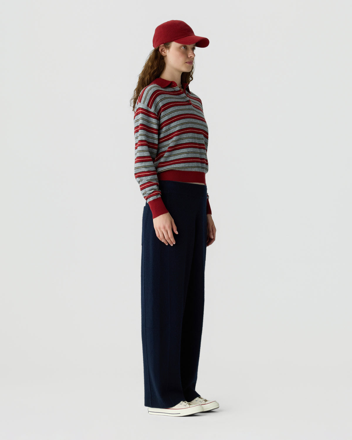 Woman wearing a striped long sleeve sweater, red baseball cap, navy wide leg trousers, and white sneakers against a light background