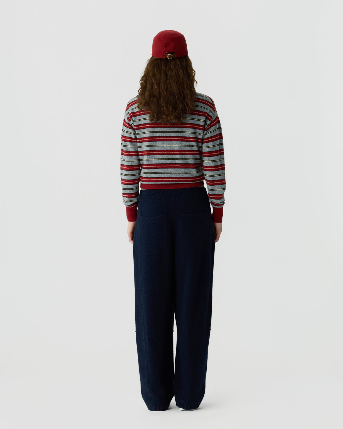 Person viewed from behind wearing a red cap a striped long sleeve sweater and wide leg navy trousers against a light background
