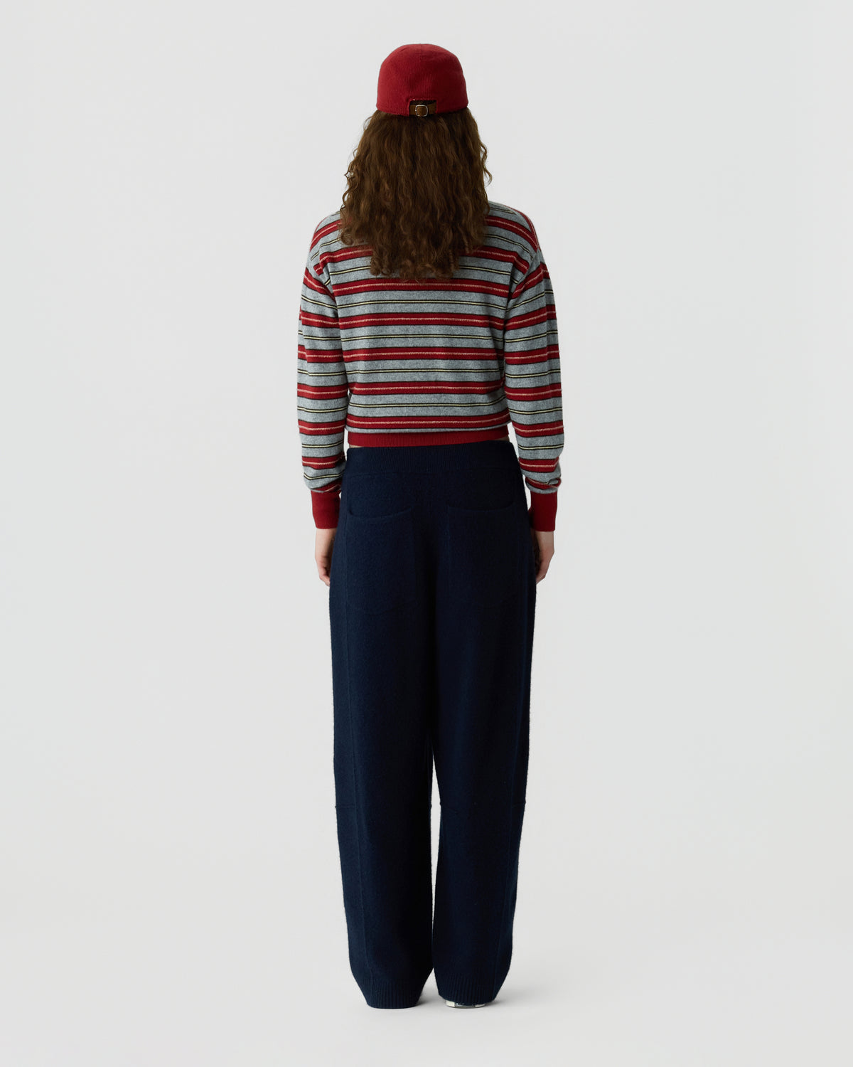 Back view of a person wearing a red hat a striped red gray and white sweater and relaxed navy trousers against a light background