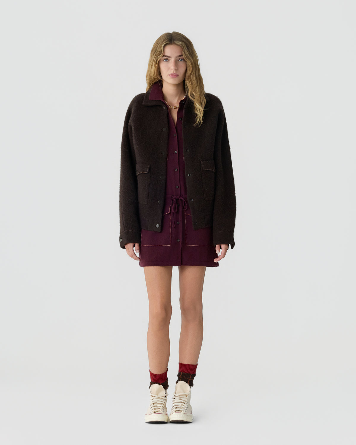Model wearing a dark brown jacket over a burgundy mini dress with burgundy socks and cream high top sneakers against a light gray background