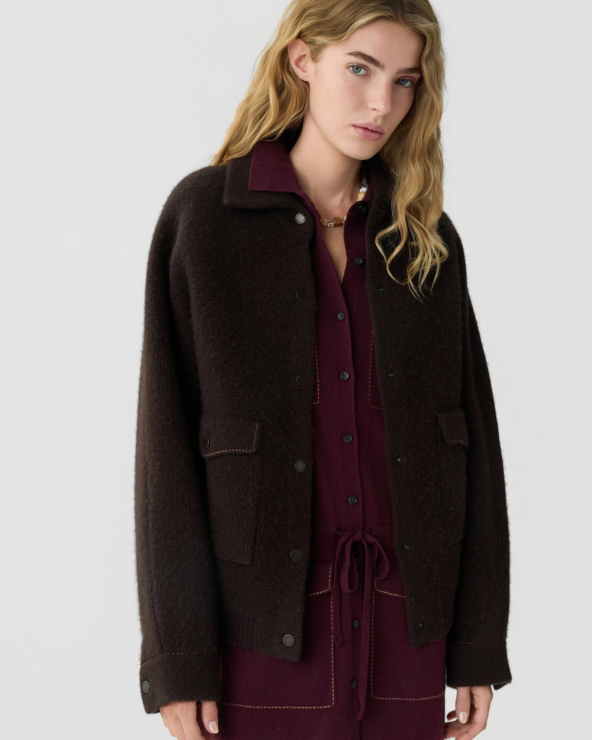 Woman wearing a dark brown textured jacket over a burgundy collared outfit against a light gray background.