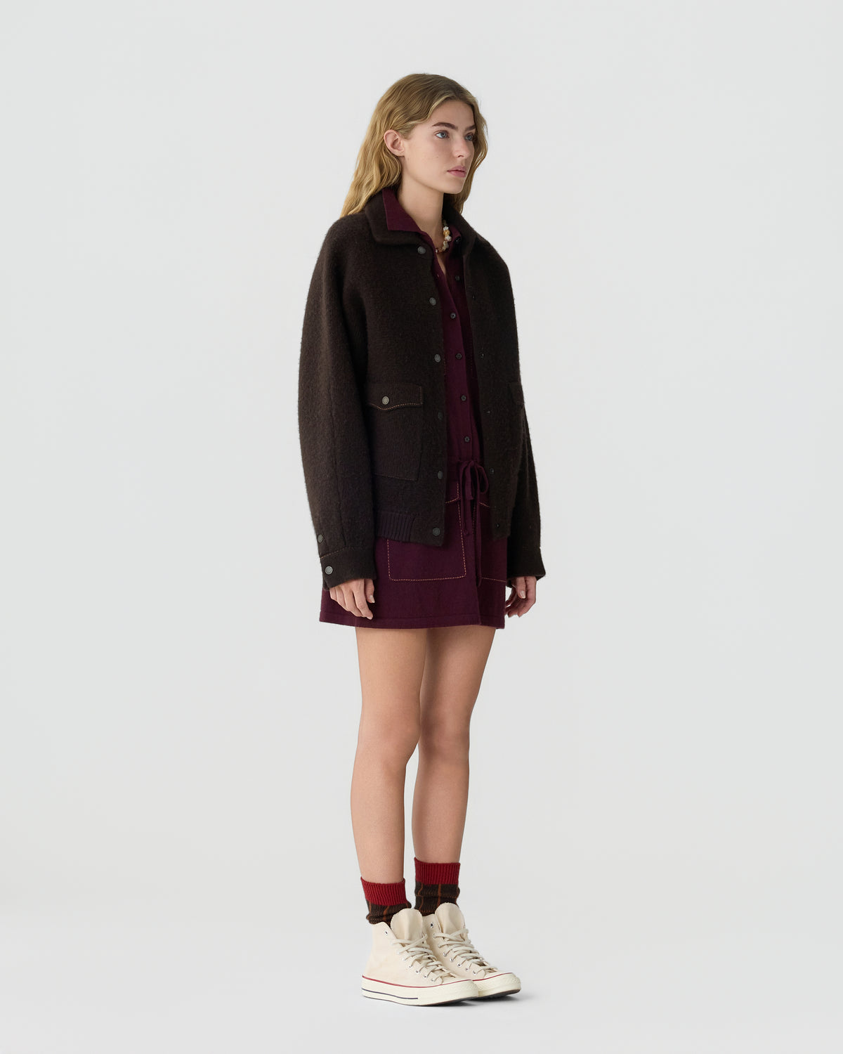 Woman wearing a dark brown jacket over a burgundy outfit with burgundy socks and white sneakers against a light gray studio background