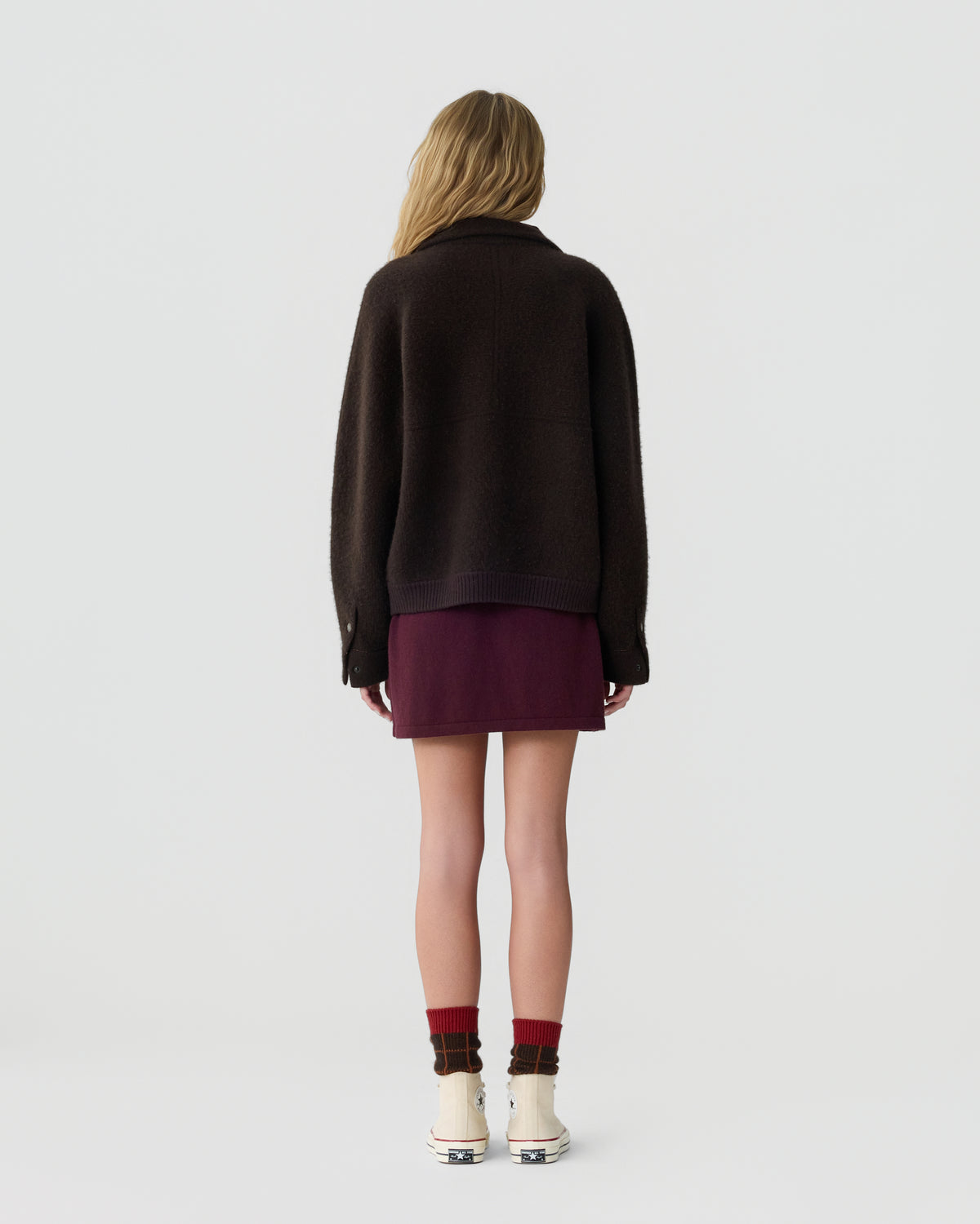 Rear view of a model wearing a dark brown sweater over a burgundy skirt with burgundy socks and light colored shoes against a plain gray background