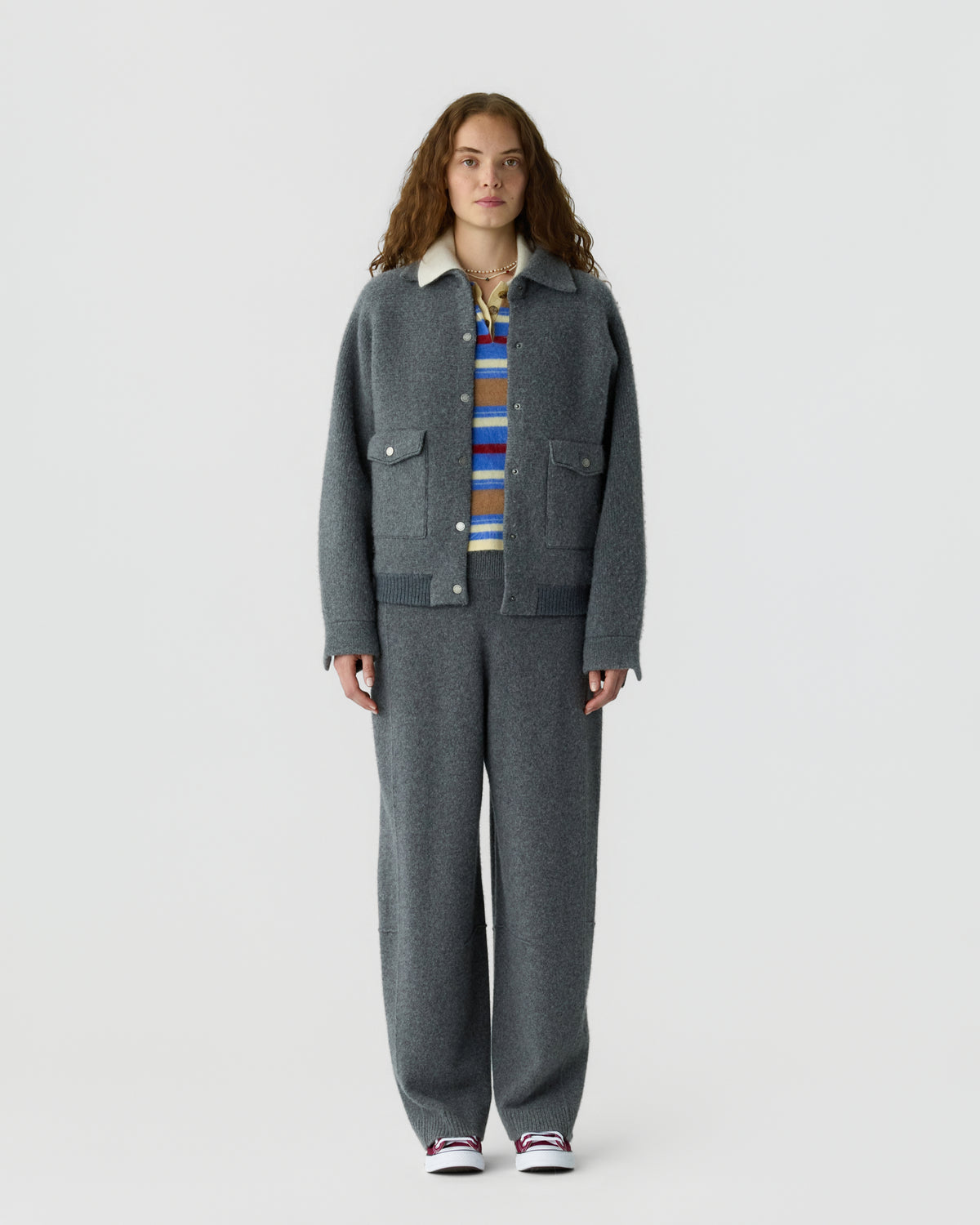 Full length portrait of a person wearing a charcoal gray collared jacket and matching trousers over a colorful striped shirt with white sneakers against a light gray background