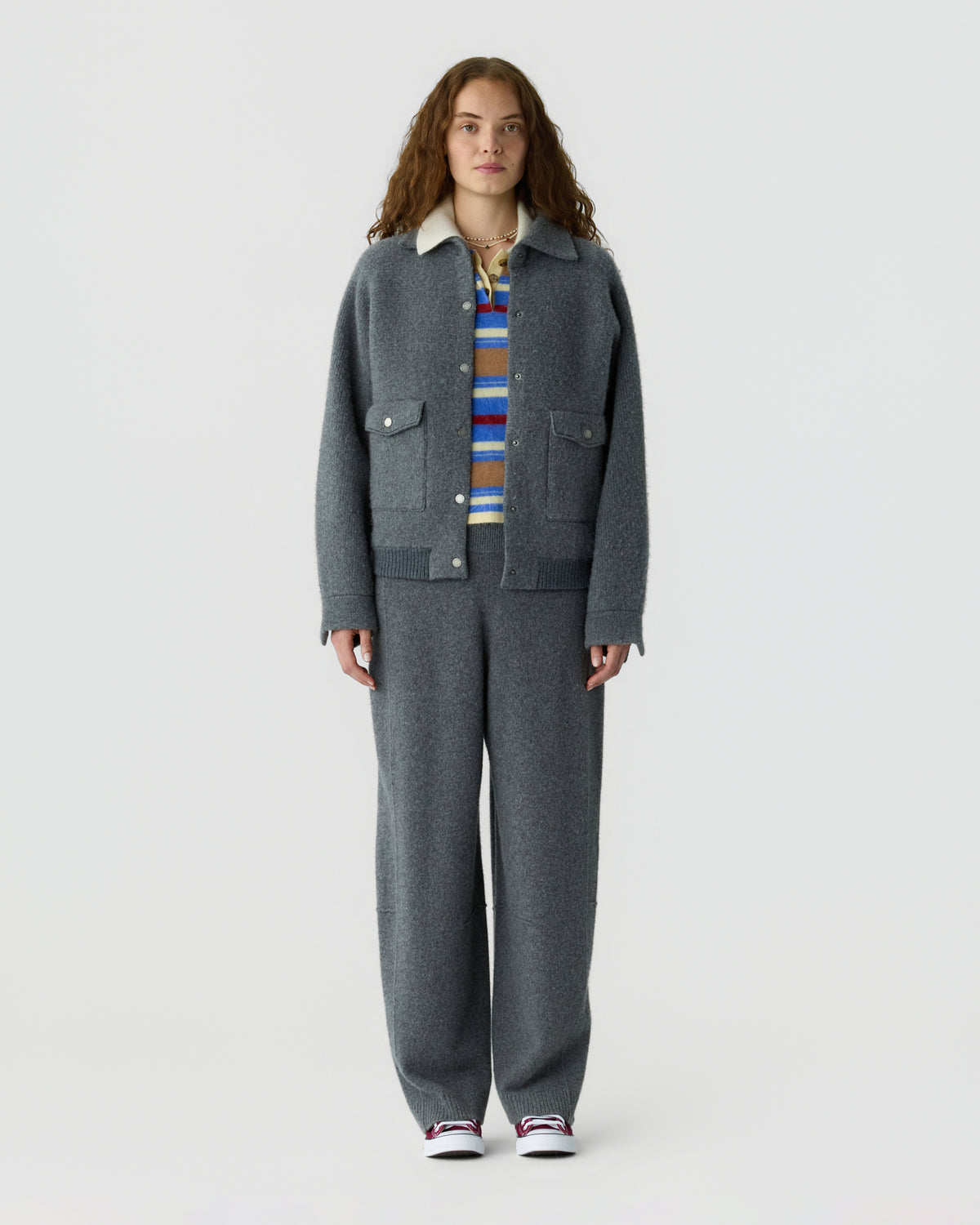 Full length model wearing a charcoal gray jacket and matching trousers over a striped sweater with white sneakers against a light gray background