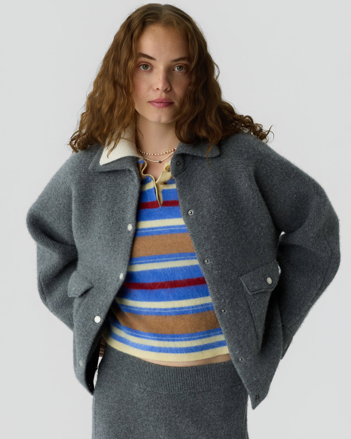 Woman with wavy hair wearing a charcoal gray buttoned jacket over a colorful striped sweater and matching gray knit skirt against a light background