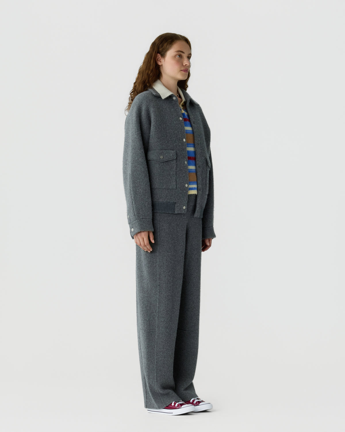 Full length view of a woman wearing a dark gray cardigan with patch pockets striped shirt wide leg trousers and white sneakers against a light background