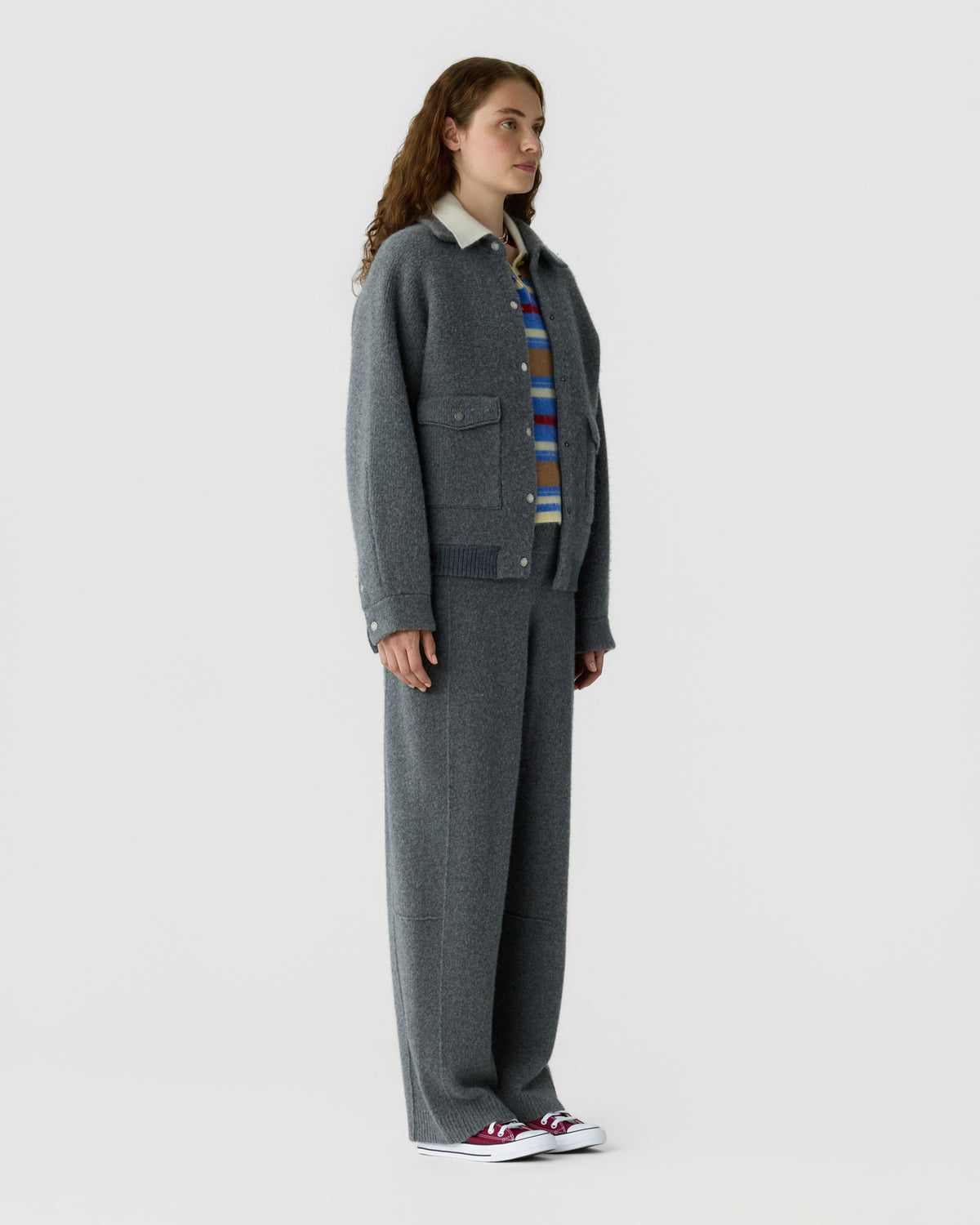 Woman wearing a charcoal gray cardigan over a striped shirt with matching wide leg trousers and red and white sneakers against a light gray background