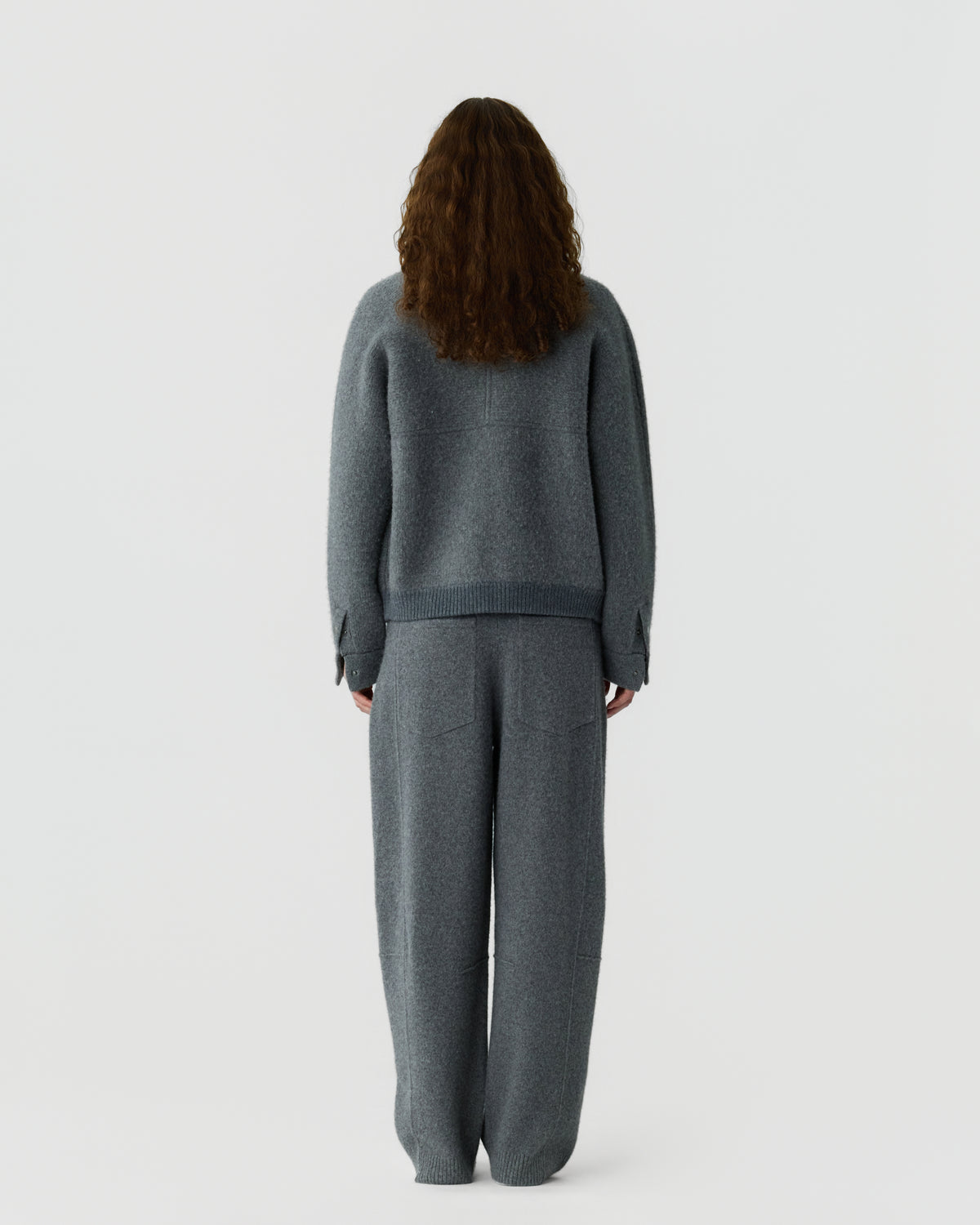 Rear view of a woman with long brown hair wearing a gray knit sweater and matching wide leg pants against a plain white background