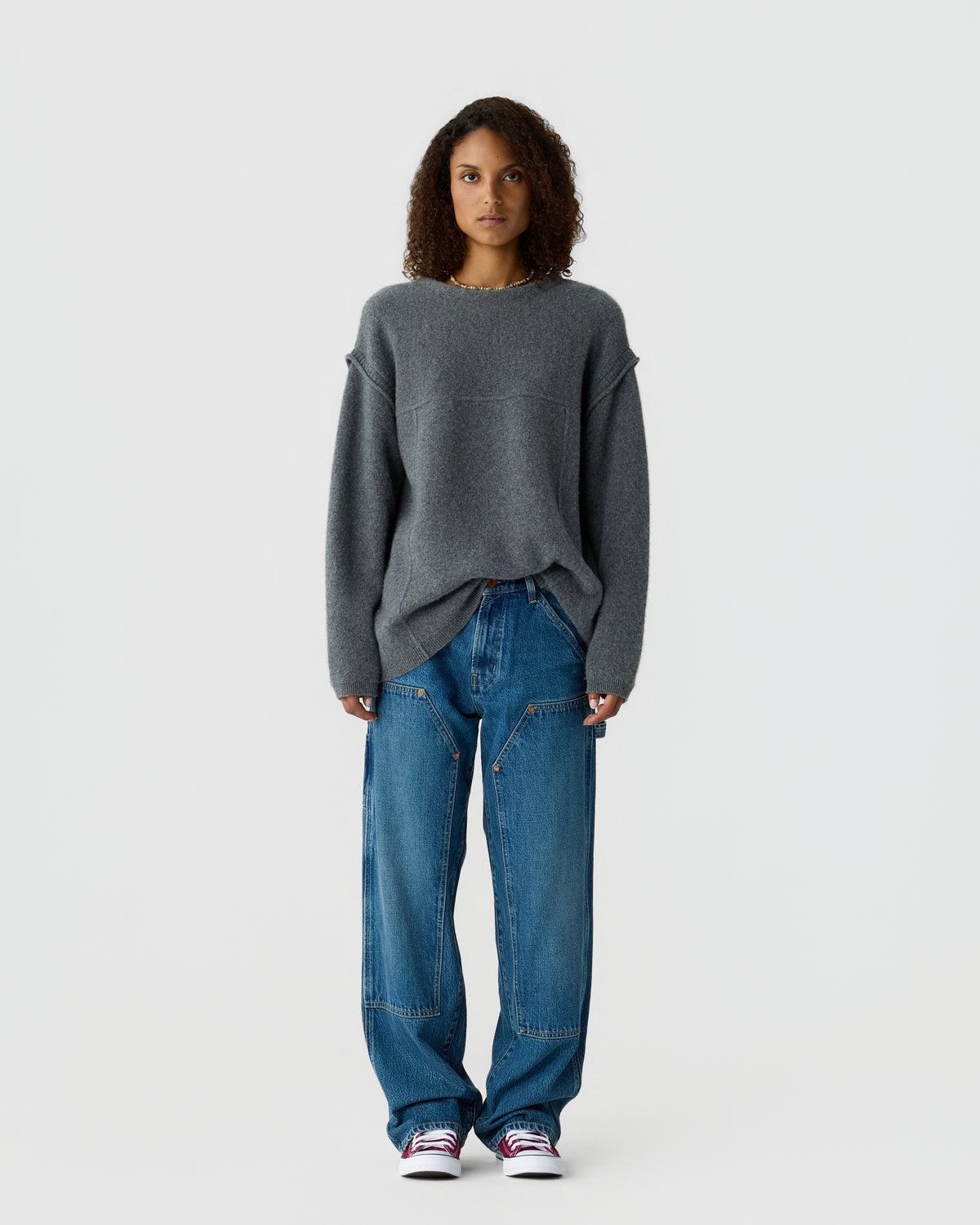 A woman with curly hair wears an oversized gray sweater and loose blue jeans while standing against a plain light gray background