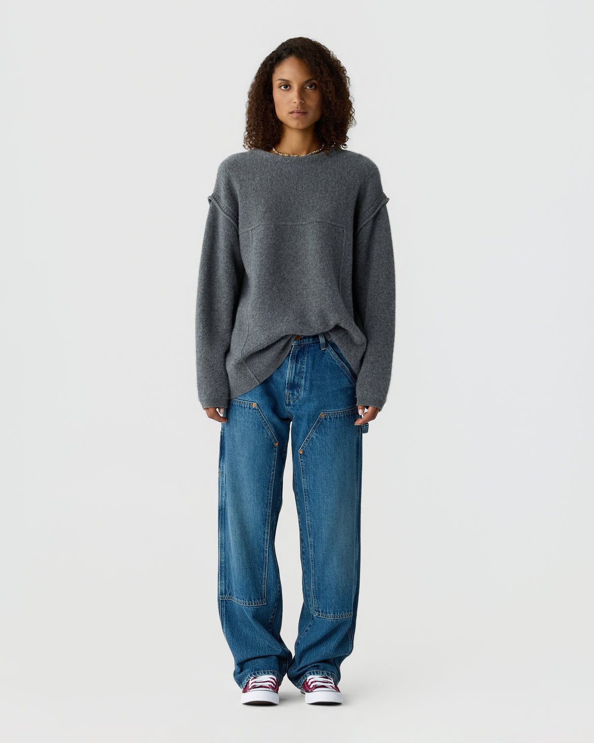 Woman standing against a light background, wearing an oversized dark gray sweater, loose blue jeans, and white sneakers with red accents