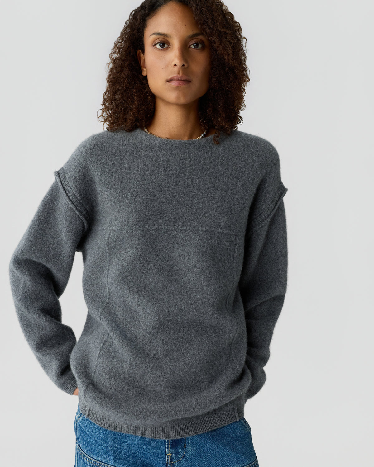 Woman with curly hair wearing a gray crew neck sweater and blue jeans against a light background