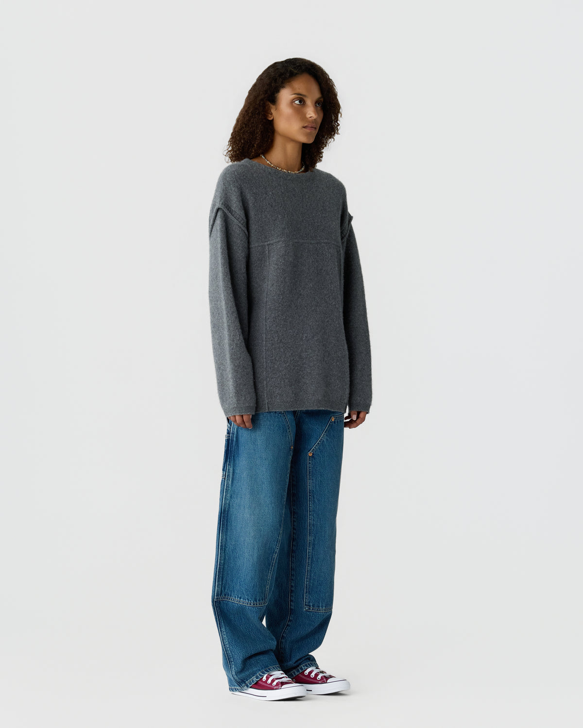 Woman wearing an oversized charcoal gray sweater with wide leg blue jeans and red and white sneakers against a light gray background