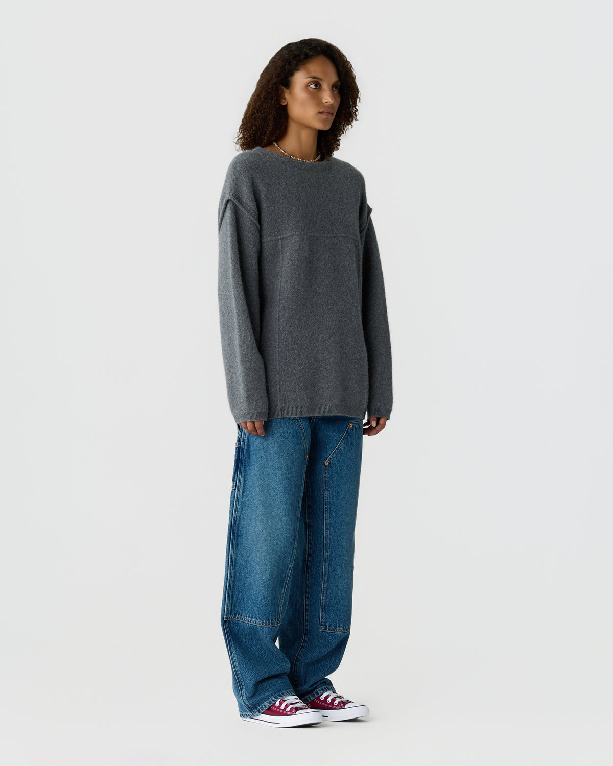 Woman wearing an oversized charcoal gray sweater, loose blue jeans, and red and white sneakers against a light gray studio background.