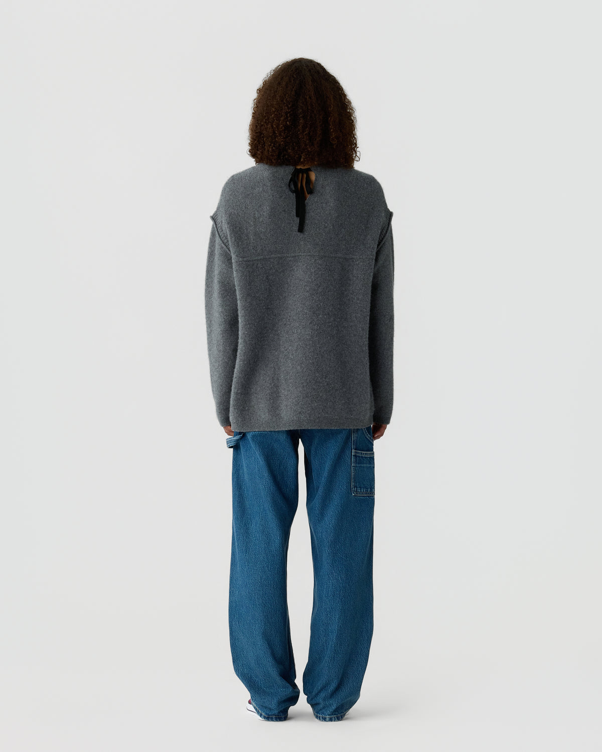 Person seen from behind wearing a charcoal gray sweater and loose blue jeans while standing against a light gray background