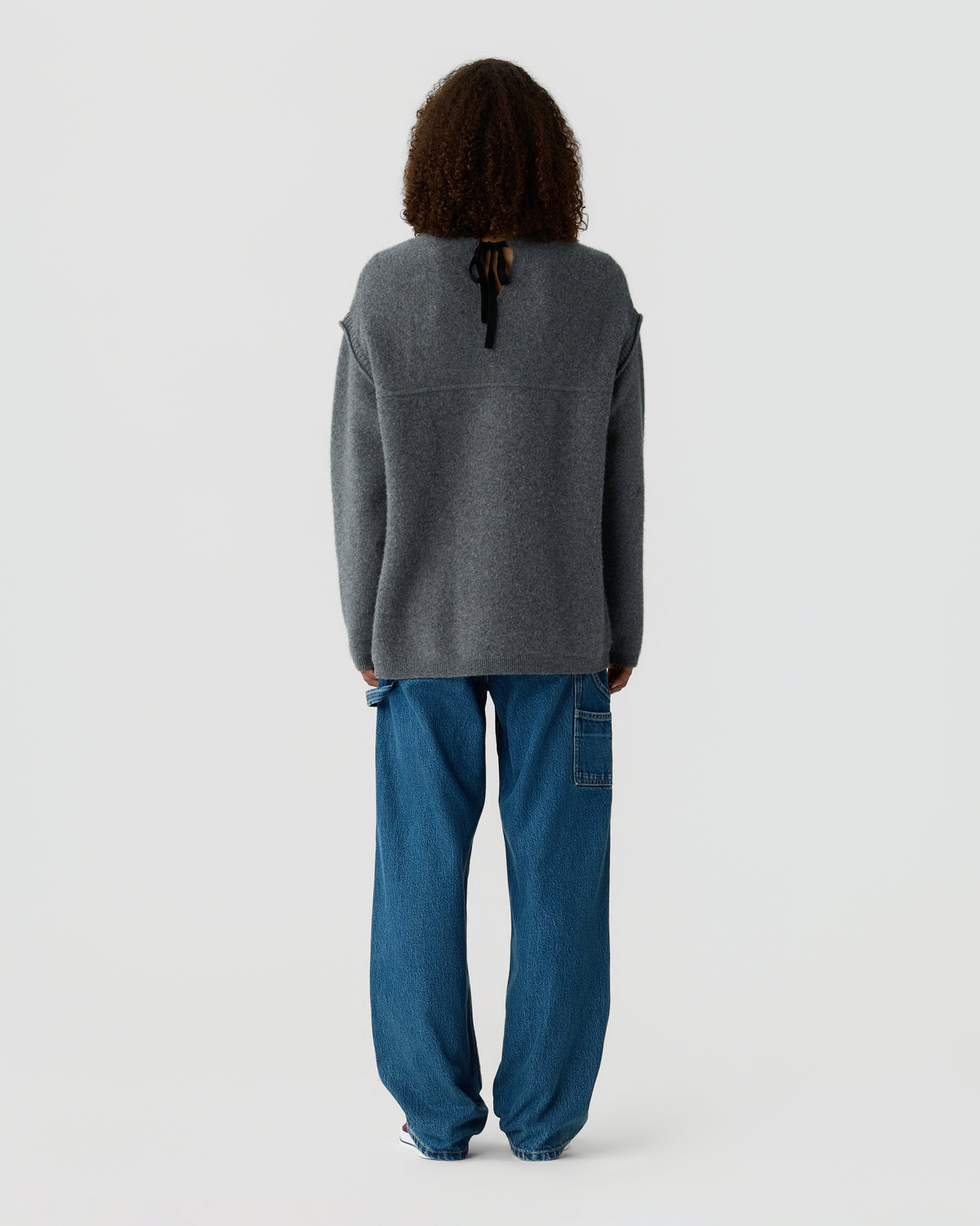 Back view of a person wearing a dark gray long sleeve sweater and loose blue jeans, standing against a plain light gray background.