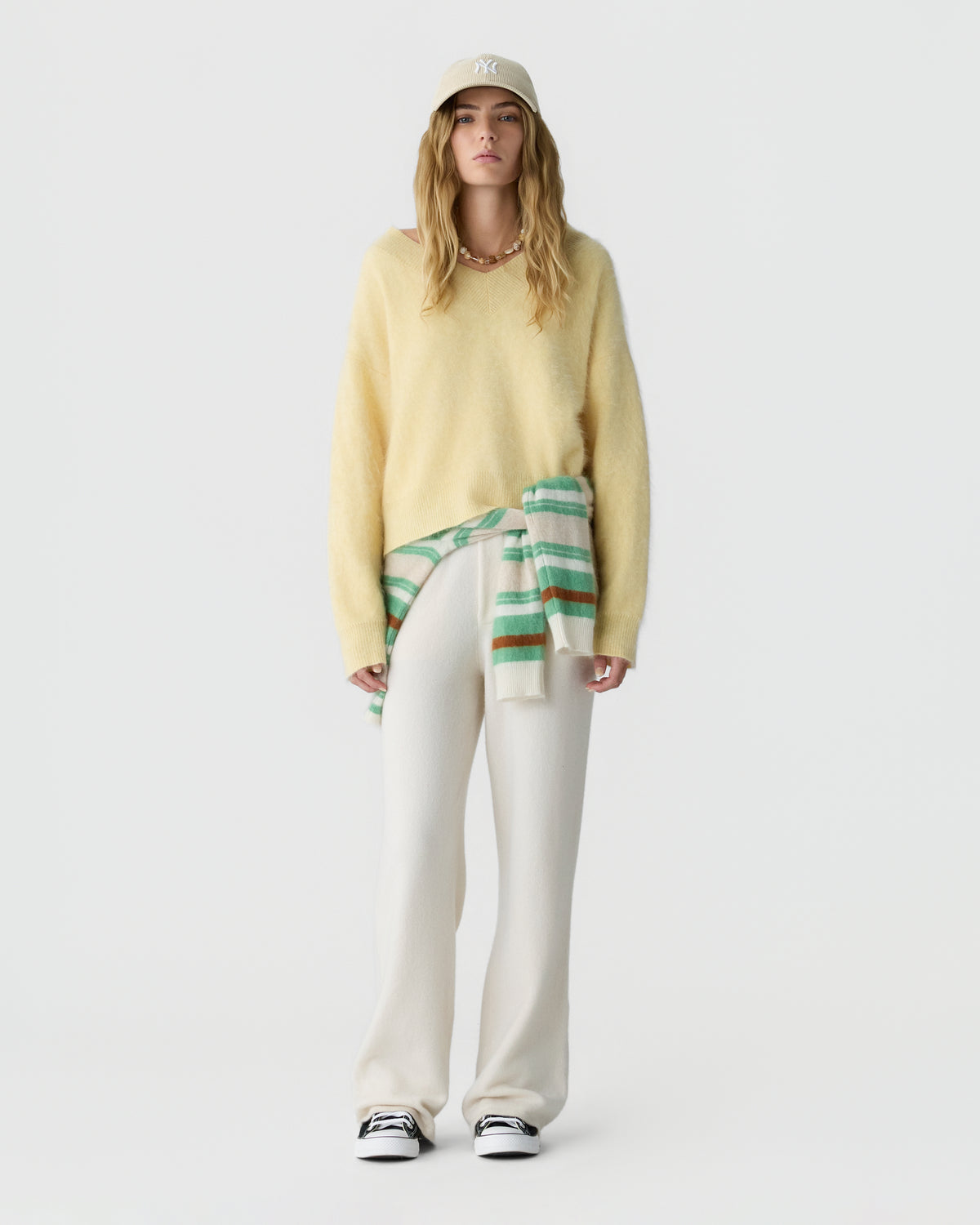 Full length view of a model wearing a pale yellow sweater, white trousers, striped green and cream scarf tied at the waist, baseball cap, and sneakers against a light background