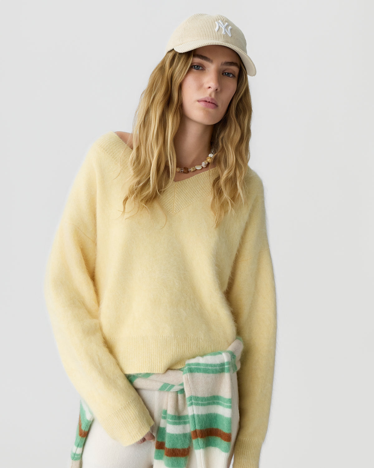 Woman with long blonde hair wearing a pale yellow sweater and beige baseball cap, with a green and cream striped garment tied around her waist.