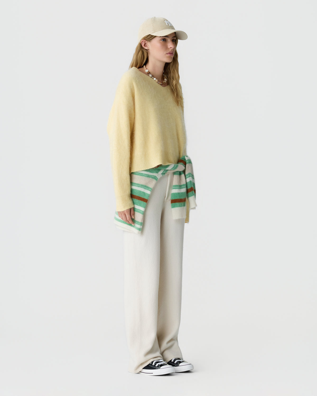 Model wearing a pale yellow sweater with a beige cap and striped green scarf tied at the waist over cream trousers and black and white sneakers.