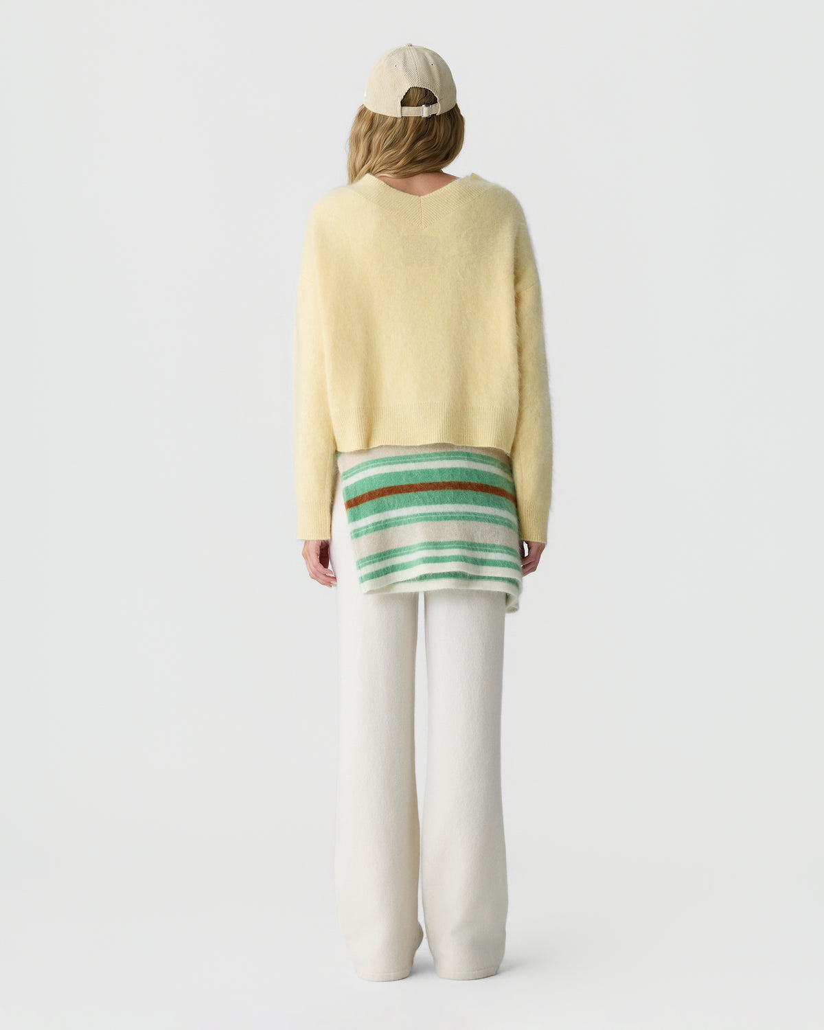 Rear view of a person wearing a pale yellow sweater, striped green and cream shirt, white trousers, and a light beige baseball cap against a plain background
