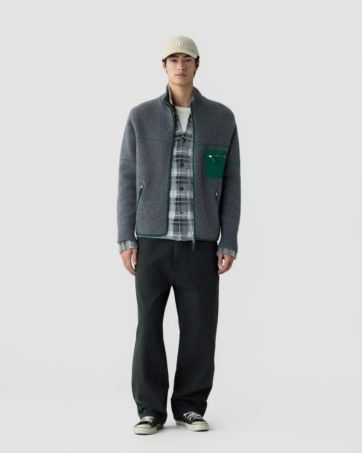 Young man wearing a grey zip-up jacket with green accents over a plaid shirt, black pants, black sneakers, and a beige baseball cap against a plain background.