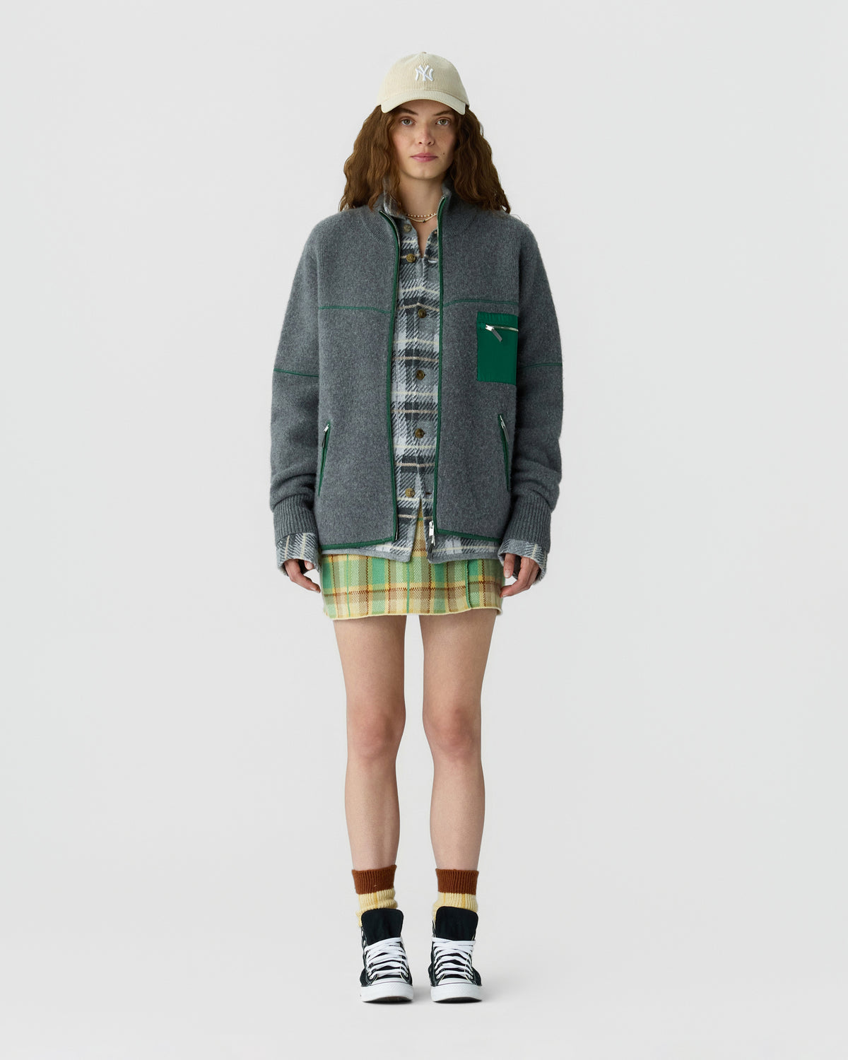 Model wearing a gray zip jacket over a plaid shirt and green yellow plaid mini skirt, styled with a baseball cap and black and white sneakers