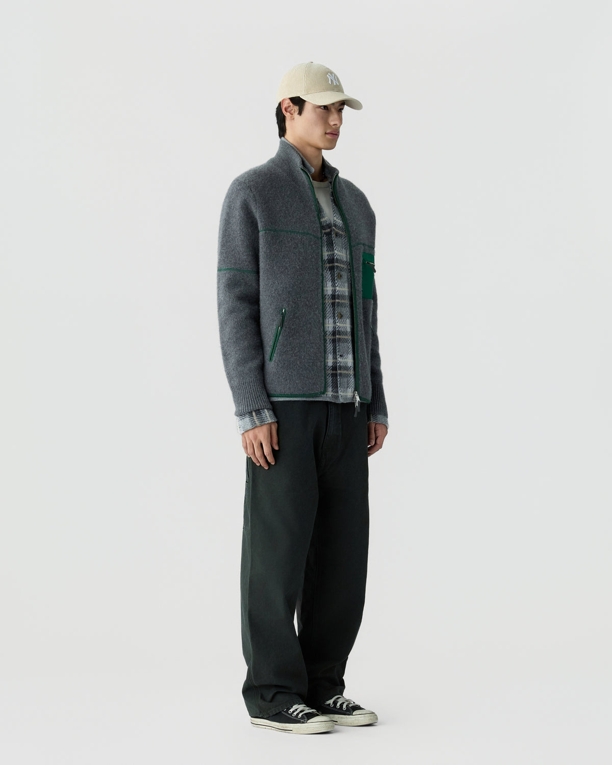 A man wearing a gray zip-up sweater layered over a gray plaid shirt, dark pants, black sneakers, and a beige baseball cap, standing against a plain white background.