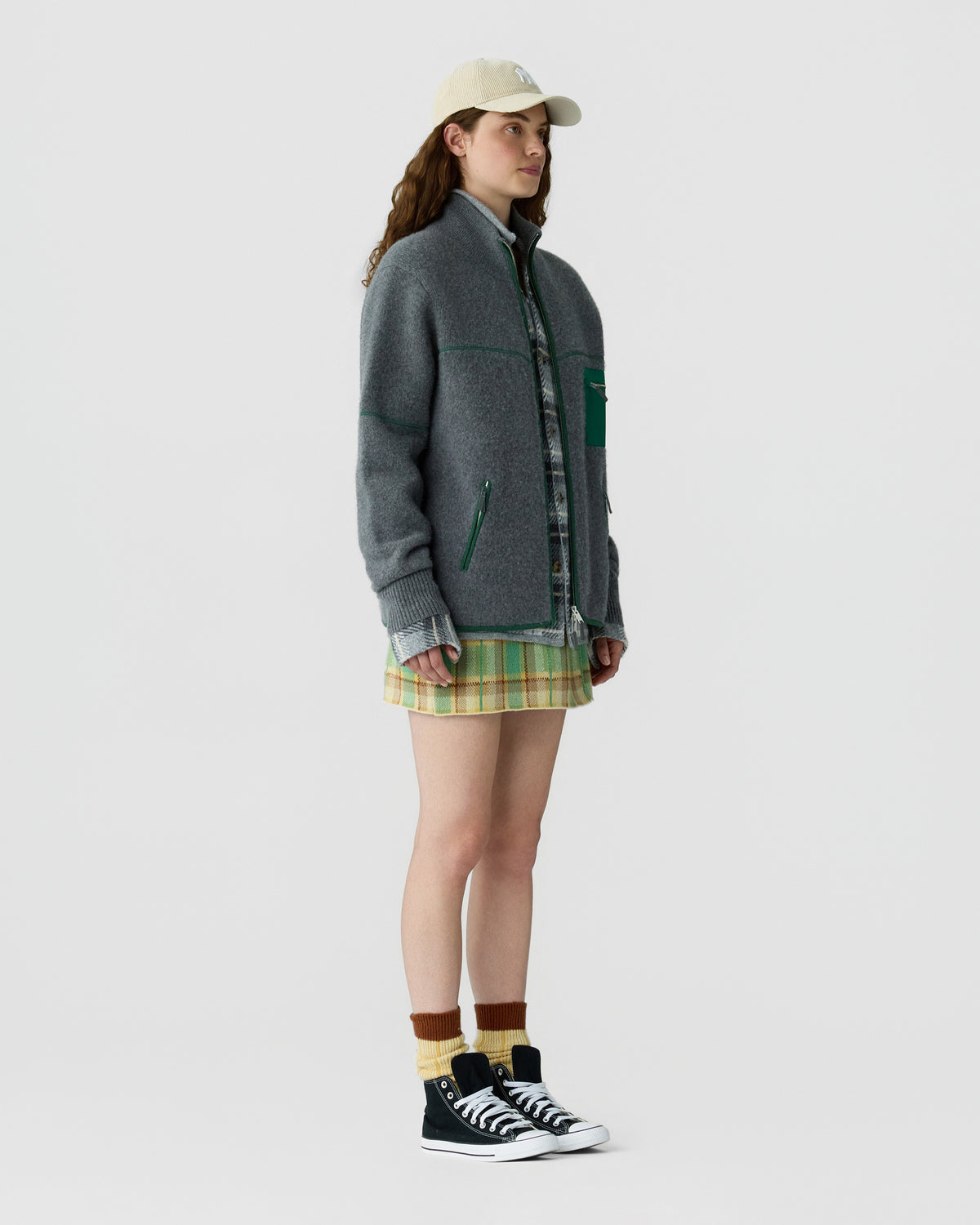Full-length view of a woman wearing a gray green zip jacket, plaid mini skirt, beige baseball cap, striped socks, and black high-top sneakers.