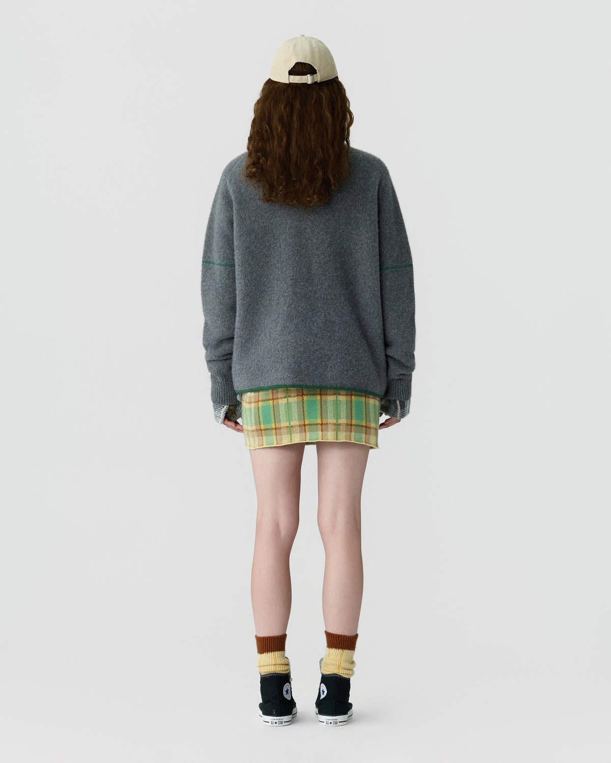 Rear view of a model wearing an oversized gray sweater over a green plaid skirt with a cream cap striped socks and black shoes against a light background