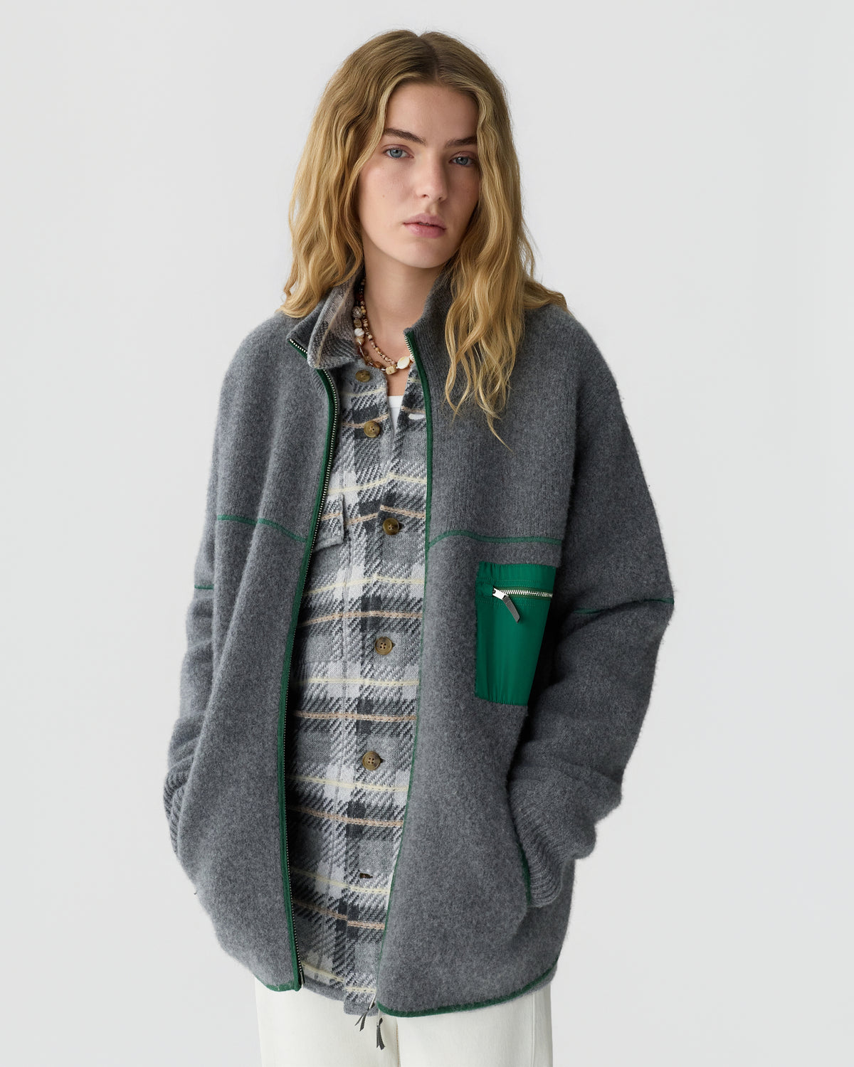 Young woman with long wavy blonde hair wearing a gray zip-up fleece jacket with green trim over a gray plaid button-up shirt, standing against a plain white background.