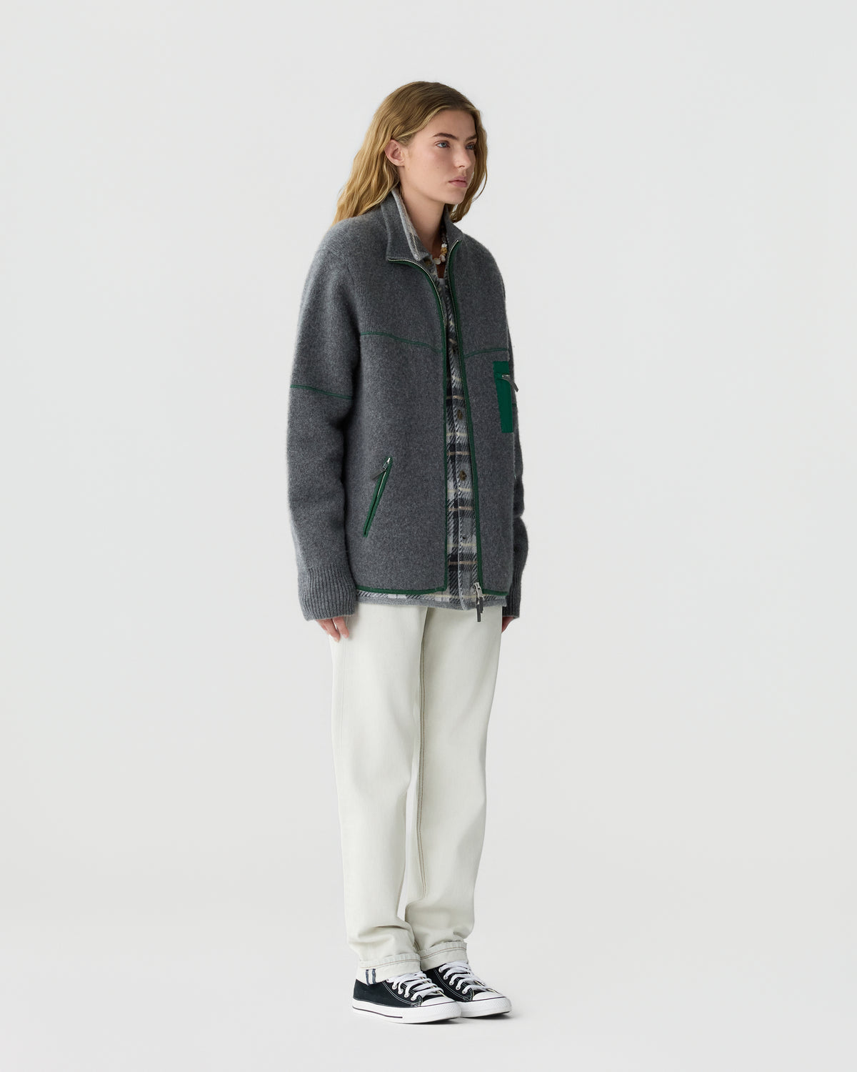 Woman standing wearing a gray zip-up jacket with green accents over a plaid shirt, white pants, and black and white sneakers against a plain light background.