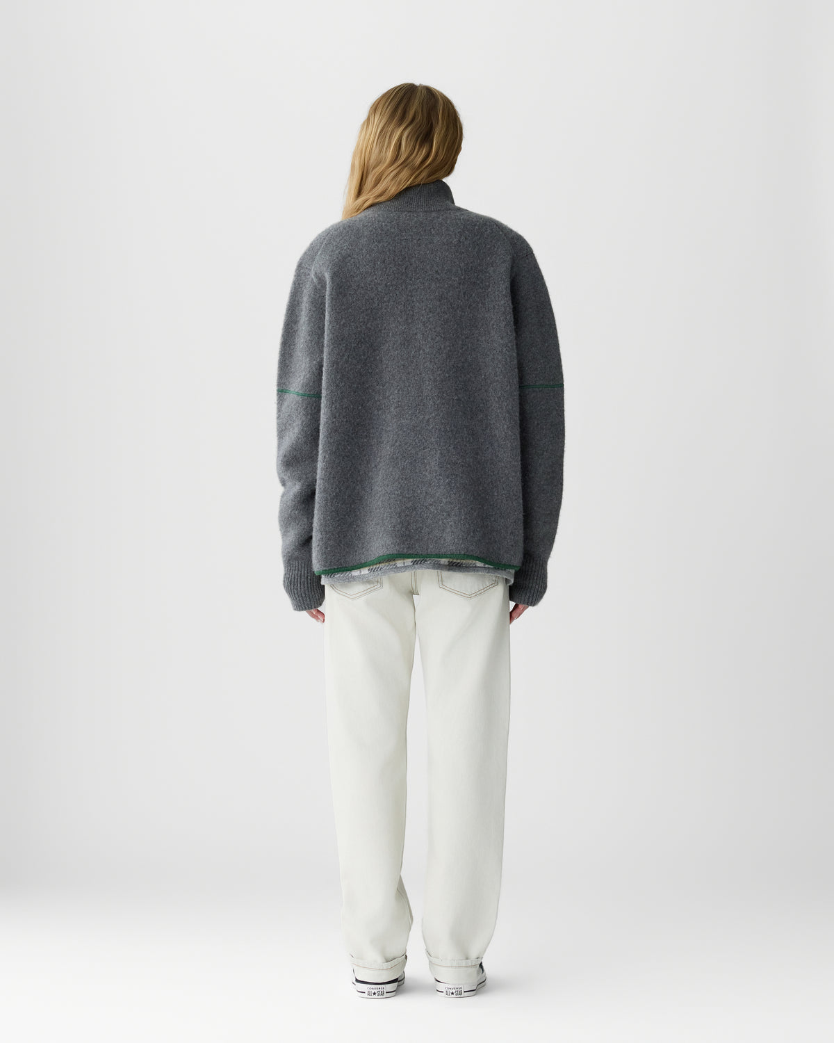 Person with long blonde hair wearing a loose dark gray sweater and white pants, standing facing away against a plain white background.