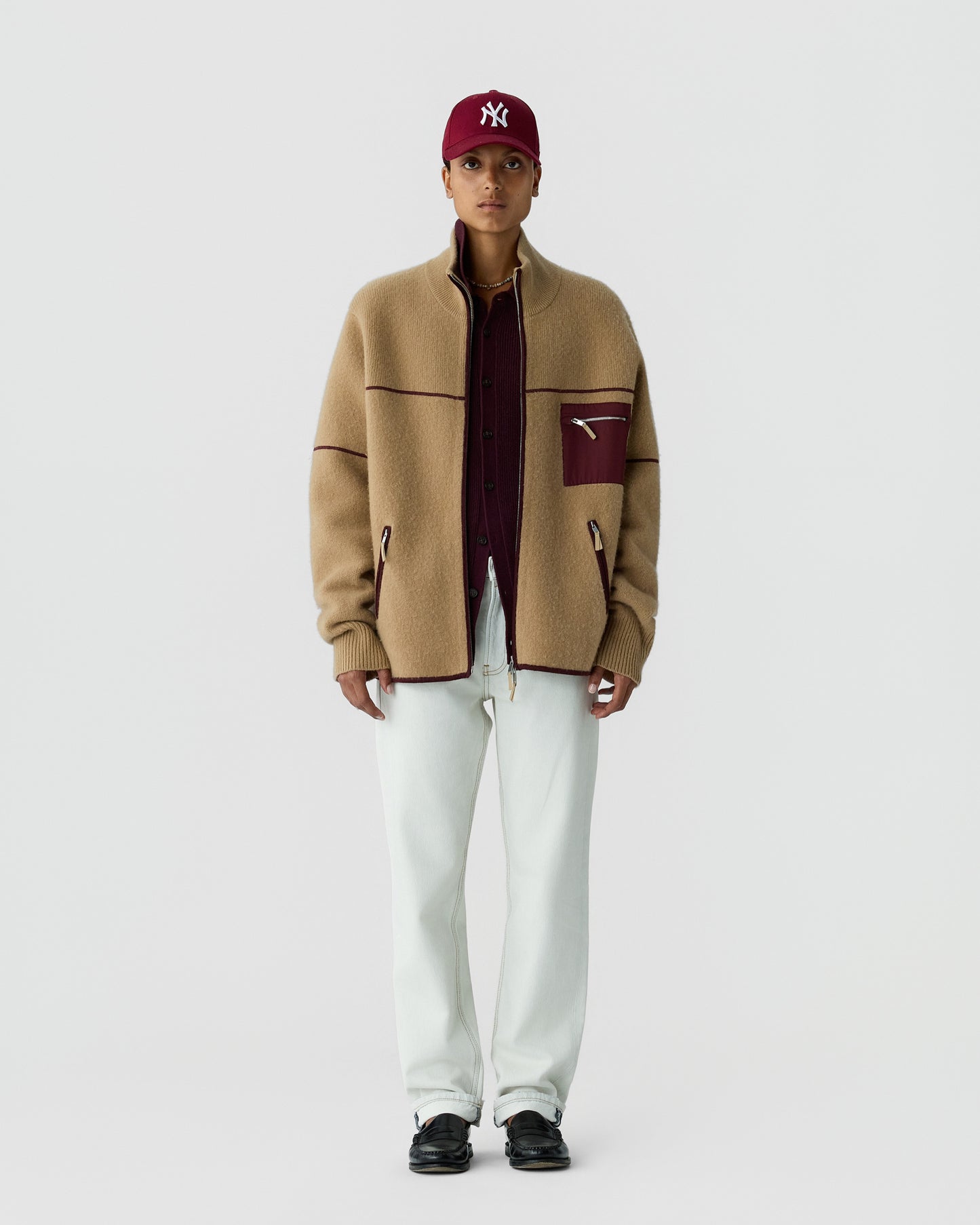 A person wearing a beige zip-up fleece jacket with maroon accents and a maroon baseball cap standing against a plain white background.
