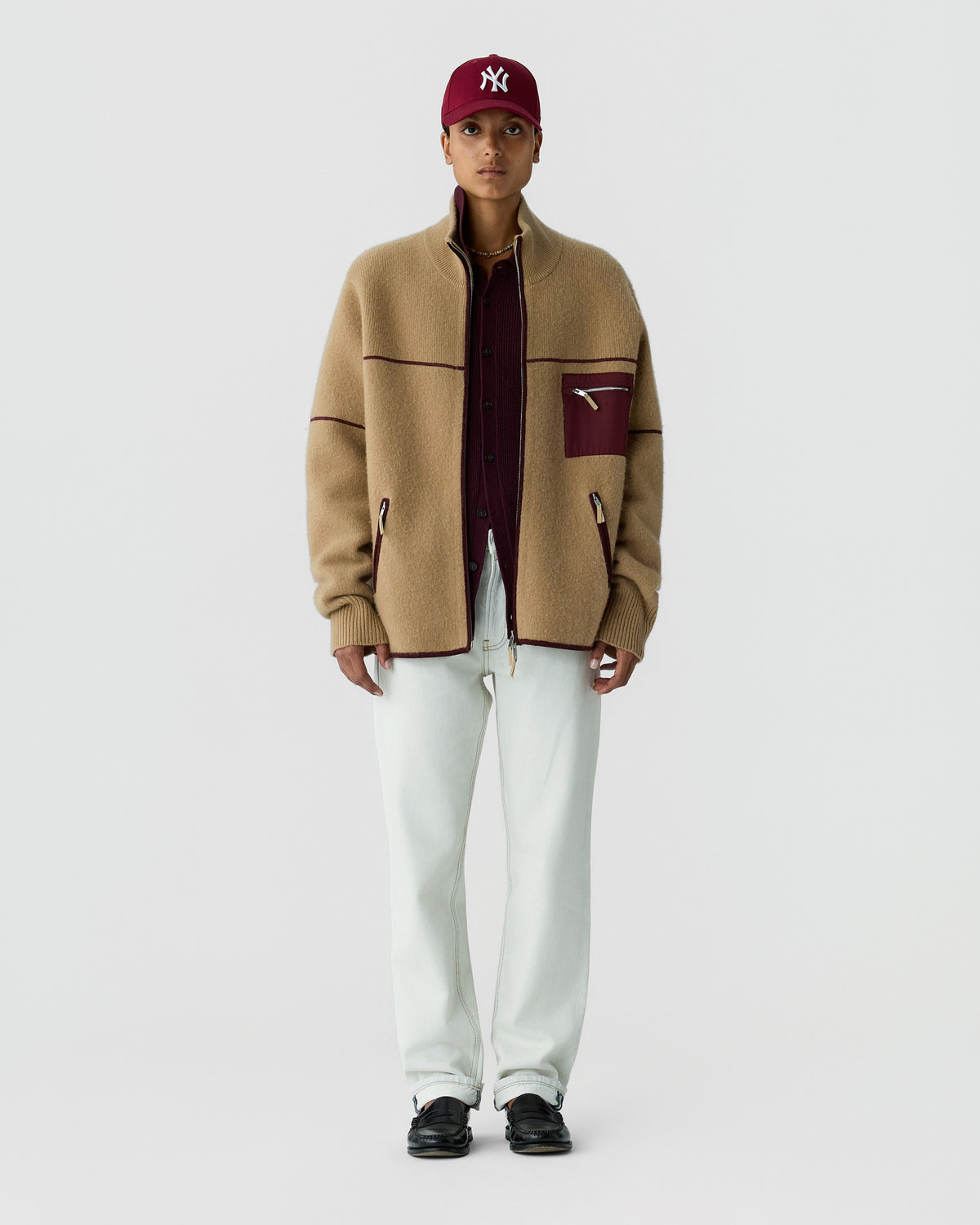 Full-length portrait of a person wearing a tan zip jacket, burgundy shirt, white trousers, black shoes, and a burgundy NY baseball cap against a light gray background.