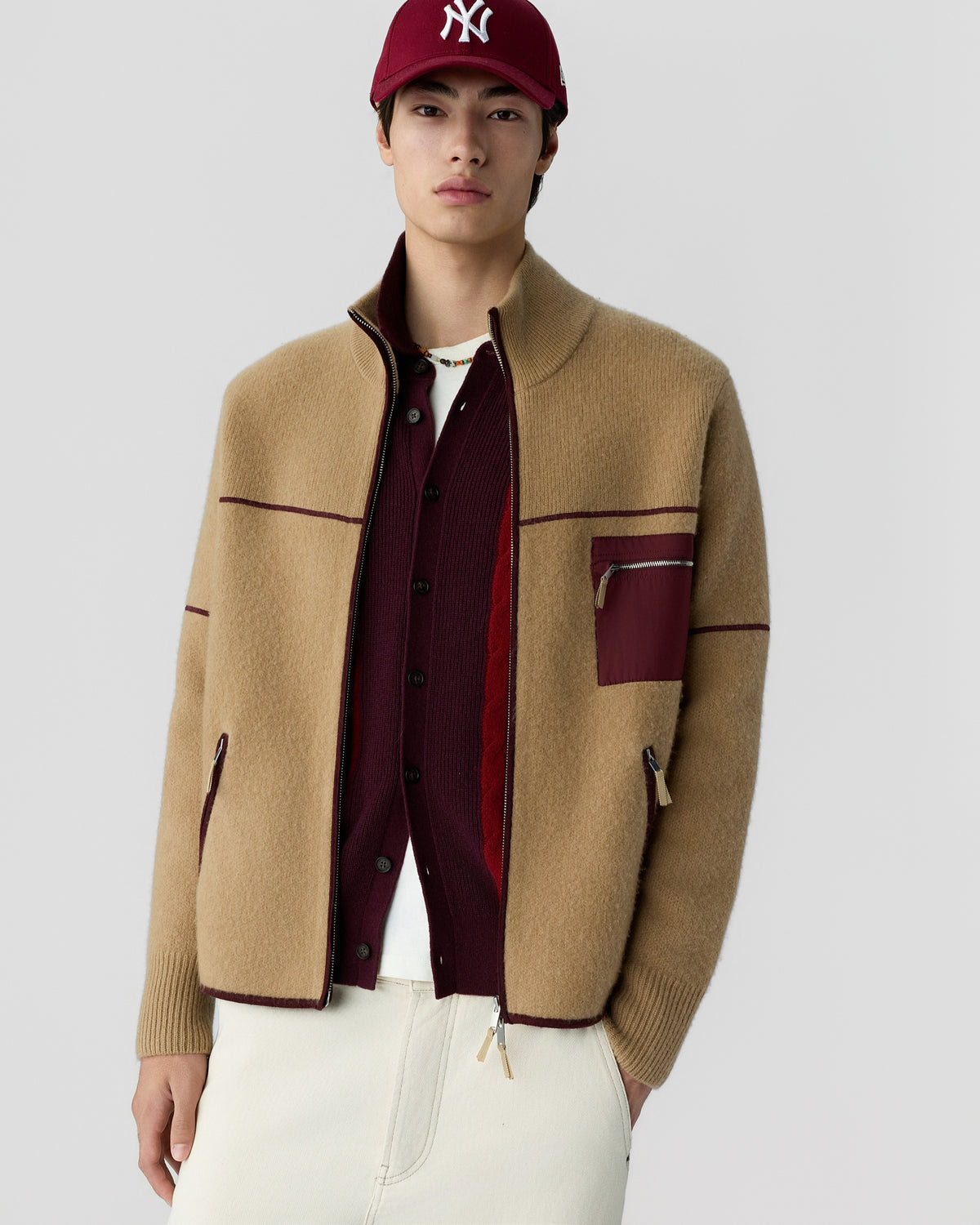 Young man wearing a tan wool jacket with burgundy accents over a burgundy button-up sweater and white pants, accessorized with a burgundy baseball cap.