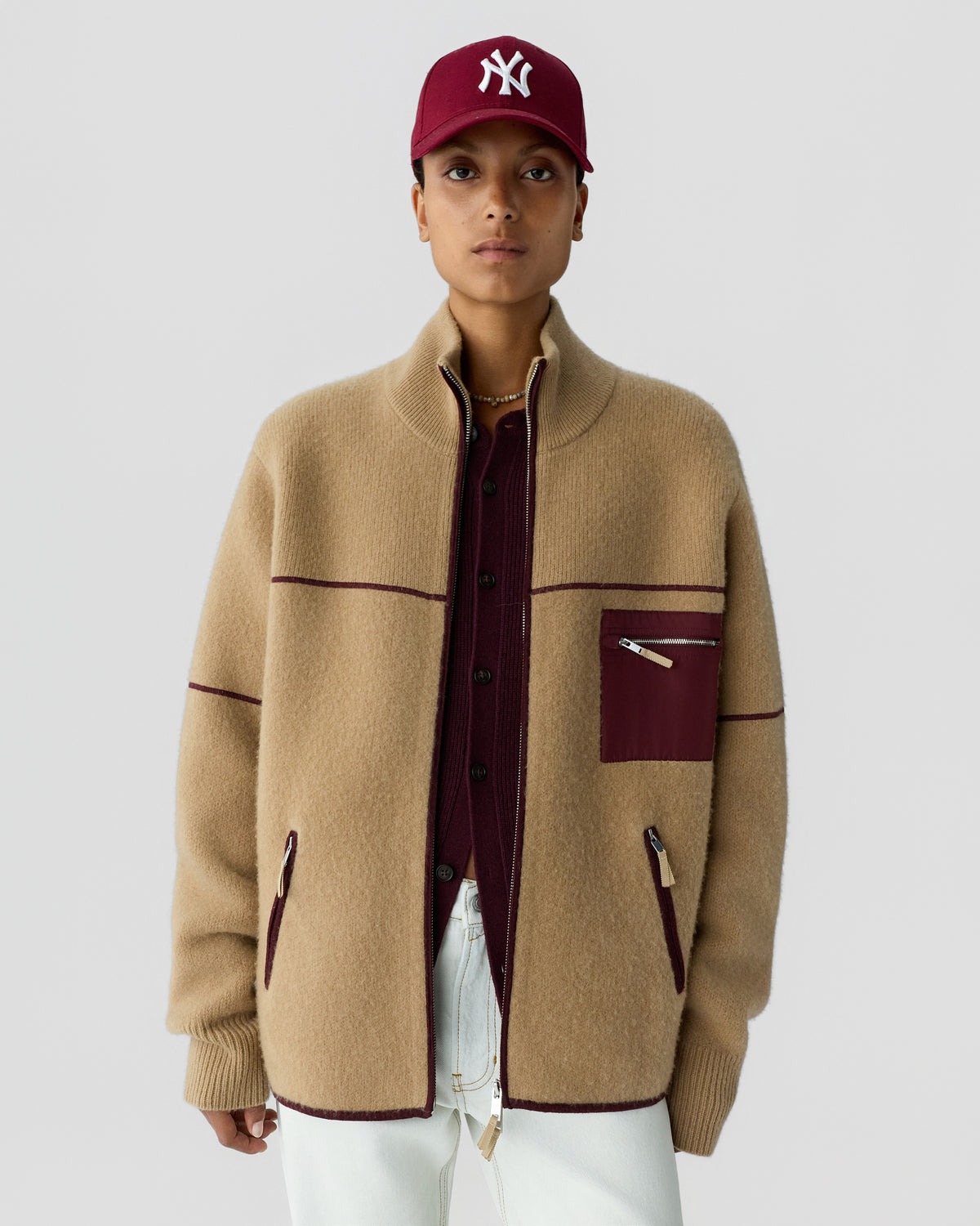 Person wearing a beige cashmere jacket with maroon trim and a maroon zippered chest pocket over a maroon buttoned shirt, white pants, and a maroon baseball cap with a white logo.